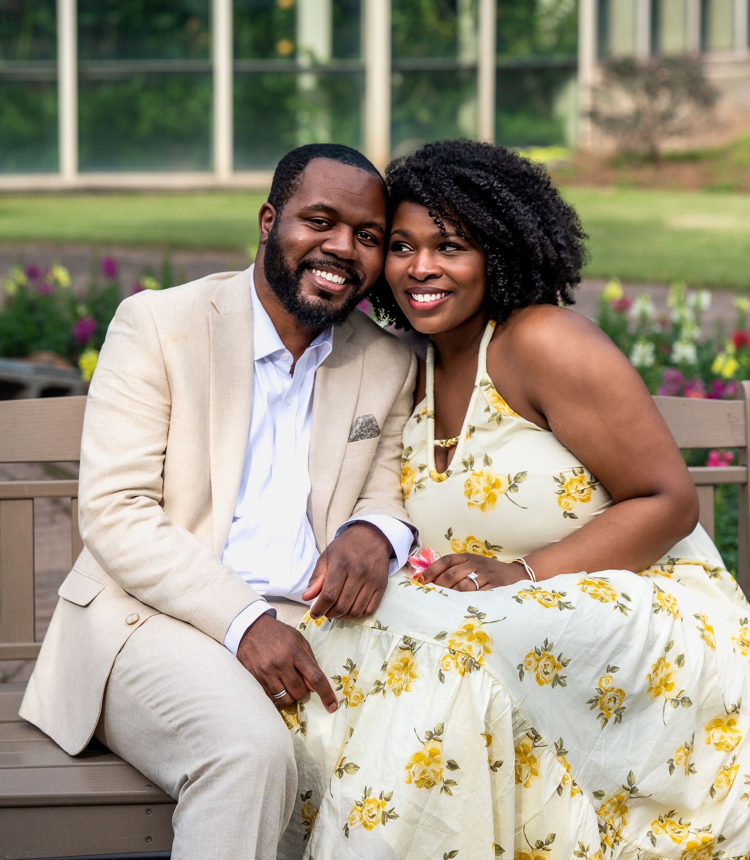 Engaged couple sitting closely on a bench smiling warmly in a sunny garden setting.