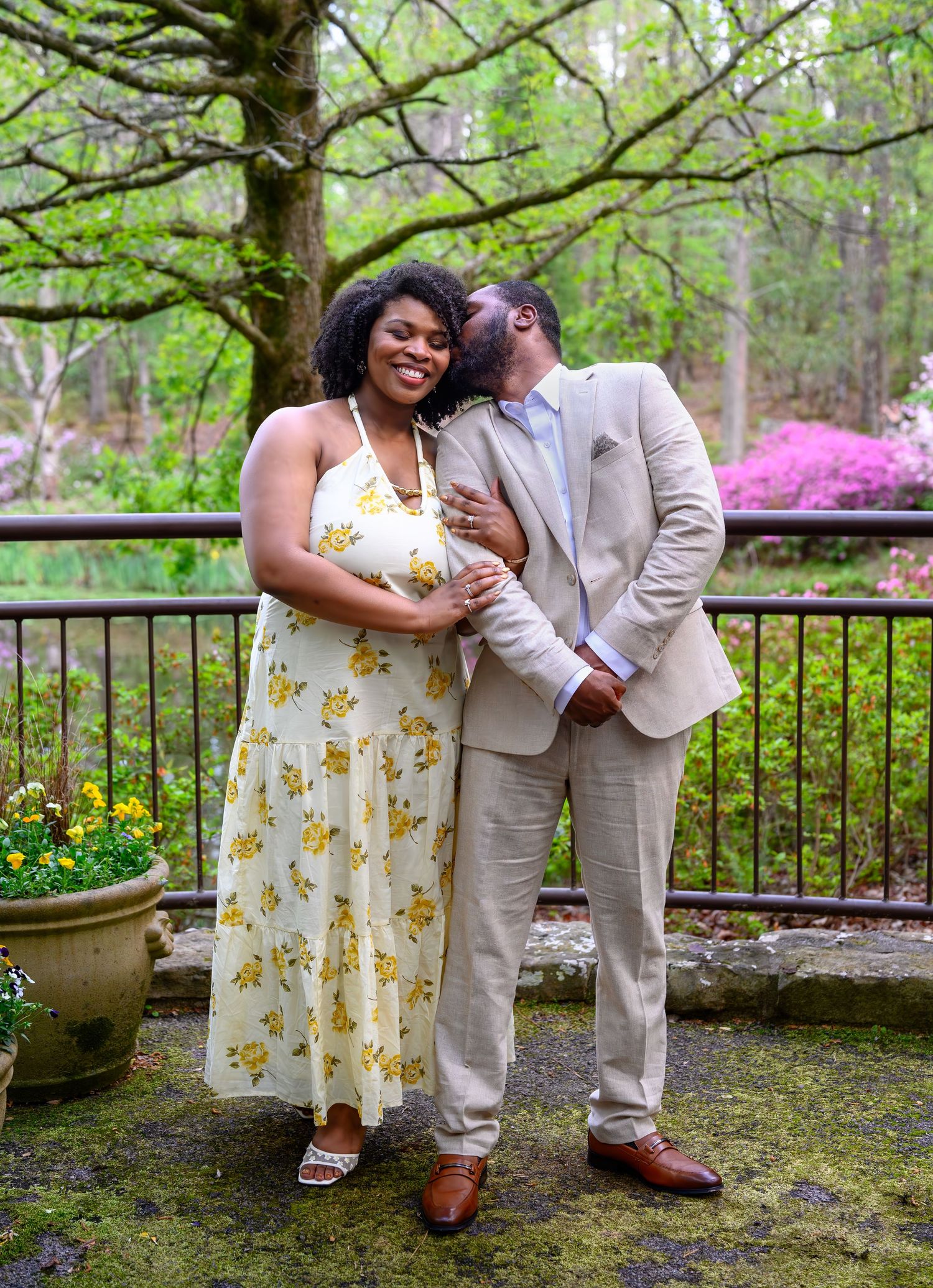 A happy couple poses together outdoors as the man kisses the womans cheek near a mossy stone terrace and trees.