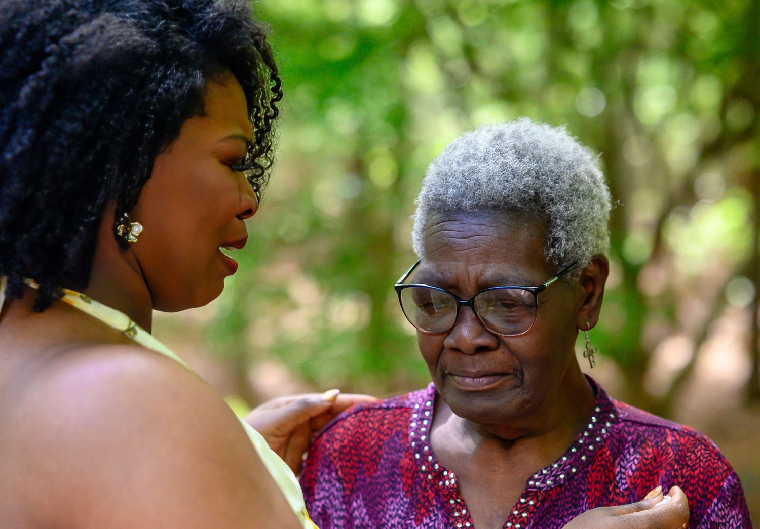 Young woman gently comforting an emotional elderly woman with short gray hair in a garden setting.