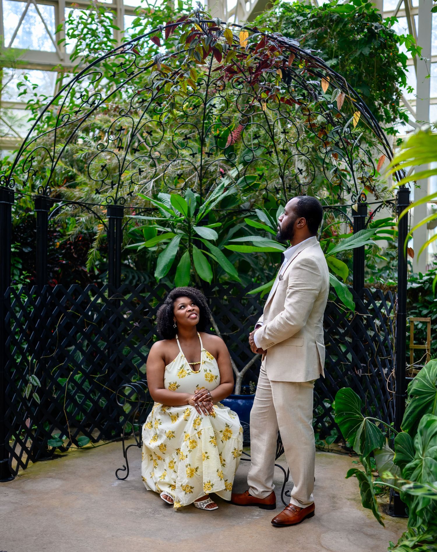 A laughing couple shares a joyful moment beneath a decorative iron arch surrounded by tropical plants indoors.