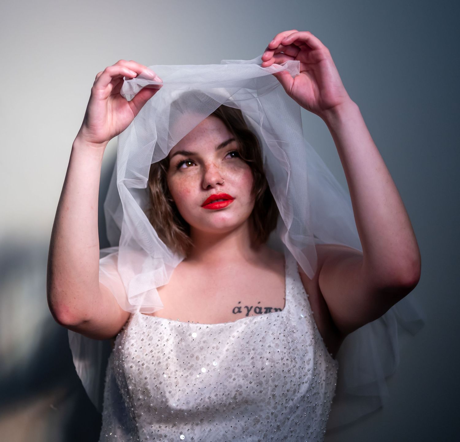 A bride in a sparkly white dress adjusts her delicate veil while posing against a moody background.