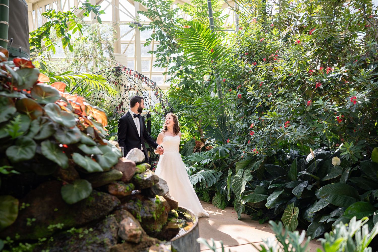 Couple in formal wedding attire poses in a lush botanical garden greenhouse setting surrounded by tropical plants.