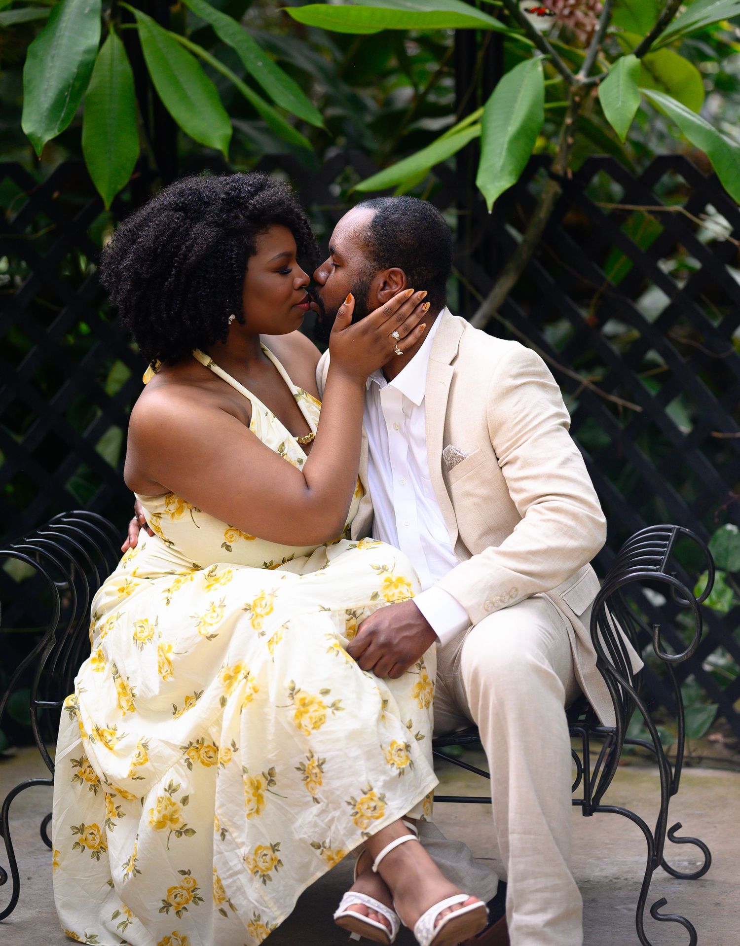 A couple shares a romantic kiss on a bench surrounded by lush green foliage in a garden.