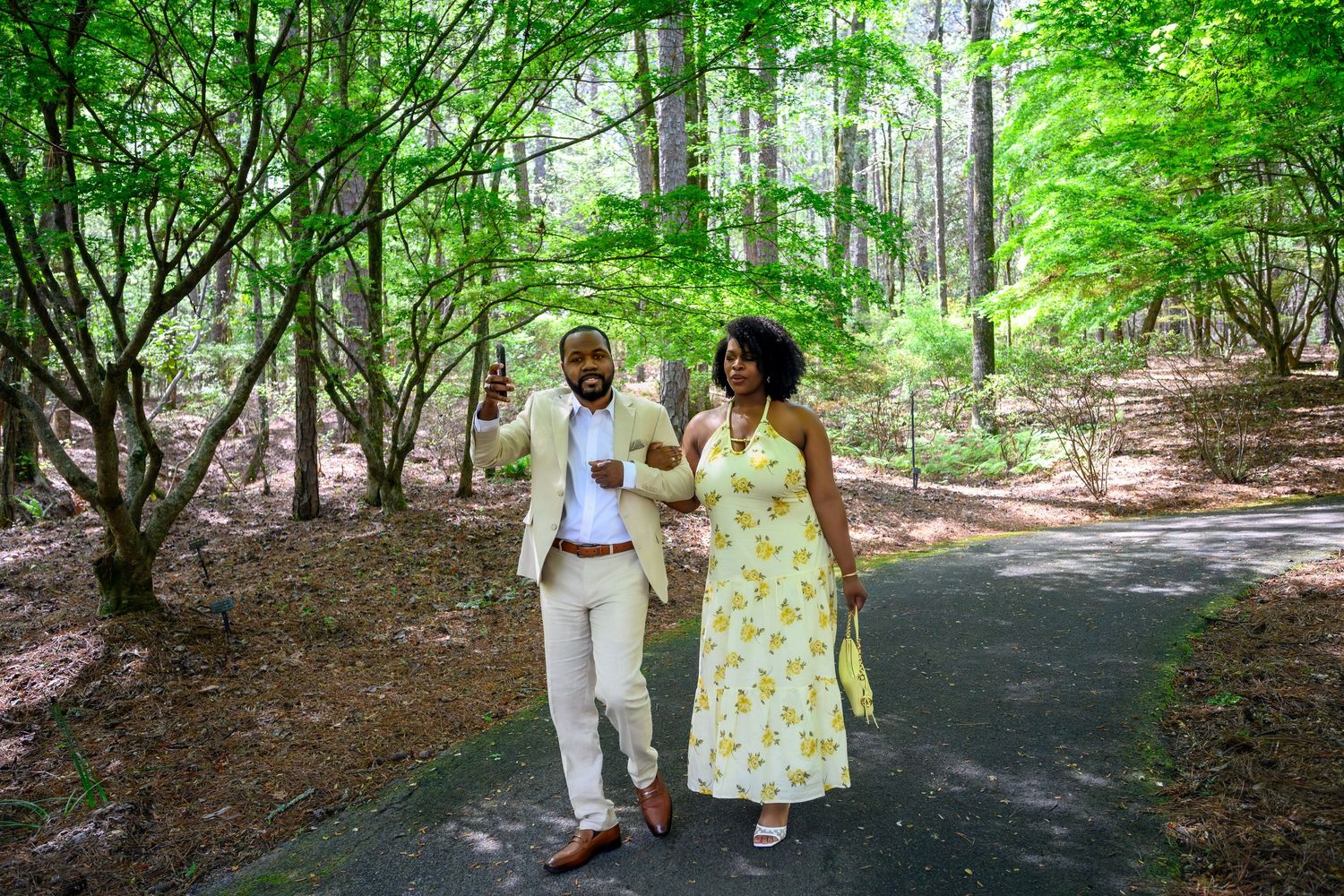 A joyful couple walks hand-in-hand on a forest path, woman in yellow floral dress, man in beige suit.