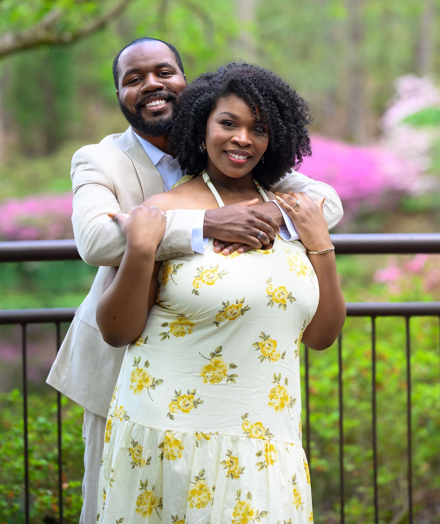 Smiling couple posing together outdoors, man in beige suit embracing woman in white yellow floral dress.