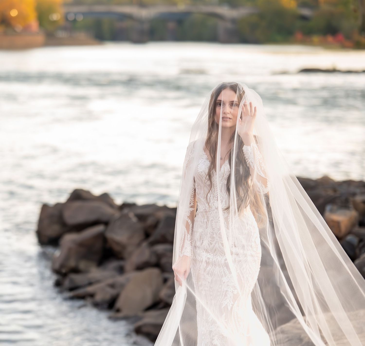 Romantic bridal photos by the water with flowing veil and lace gown at sunset.