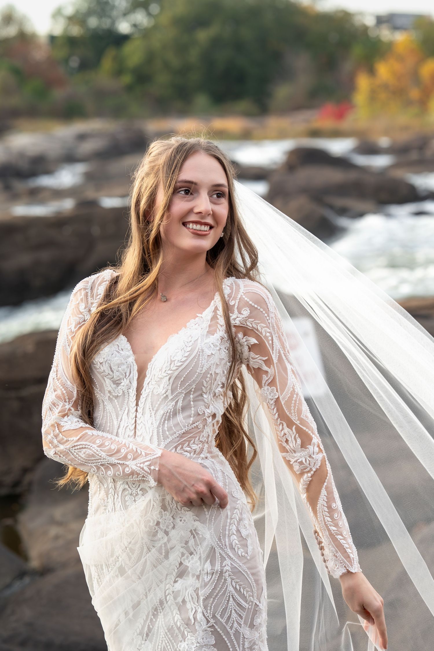 A bride in a lace wedding dress poses outdoors against rocky terrain and autumn foliage.