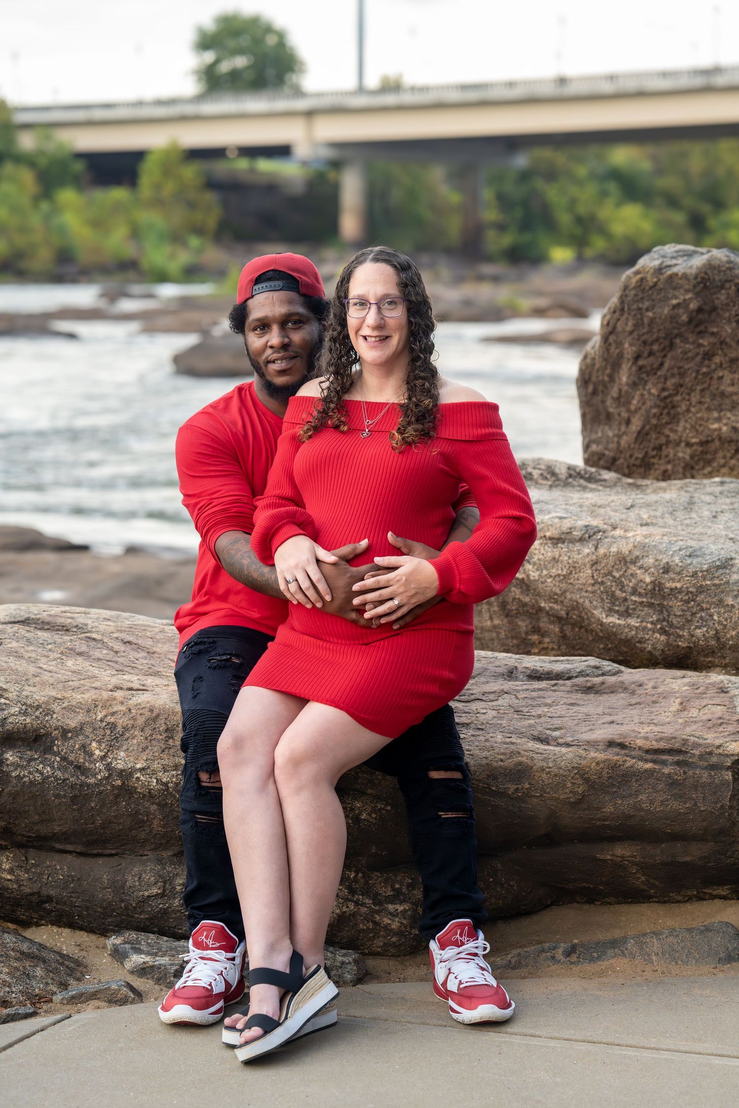 A couple in matching red sweaters sits together on large rocks by the water, showing playful and intimate poses.
