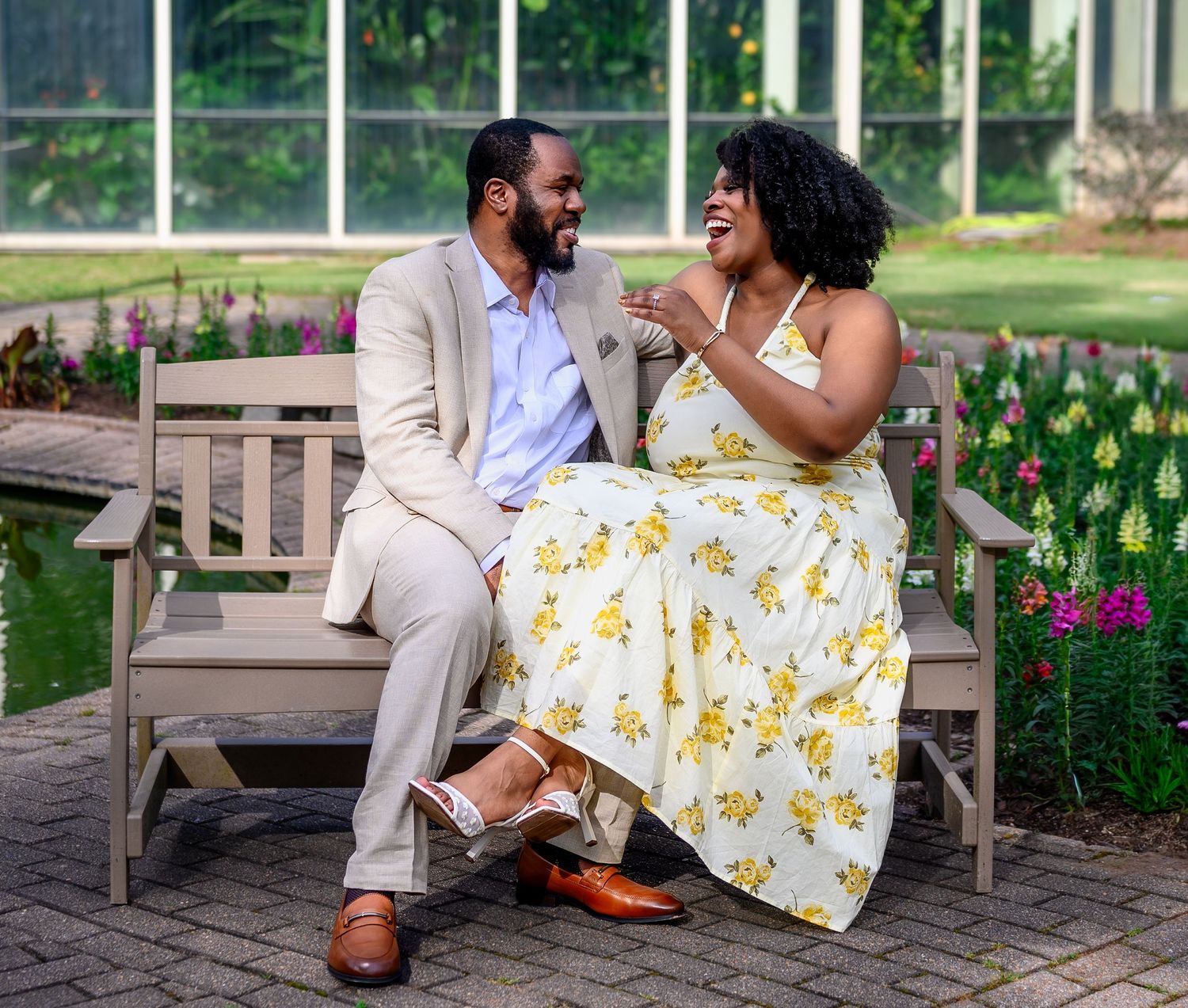 A laughing engaged couple sits together on a wooden bench in a colorful garden during their engagement session.
