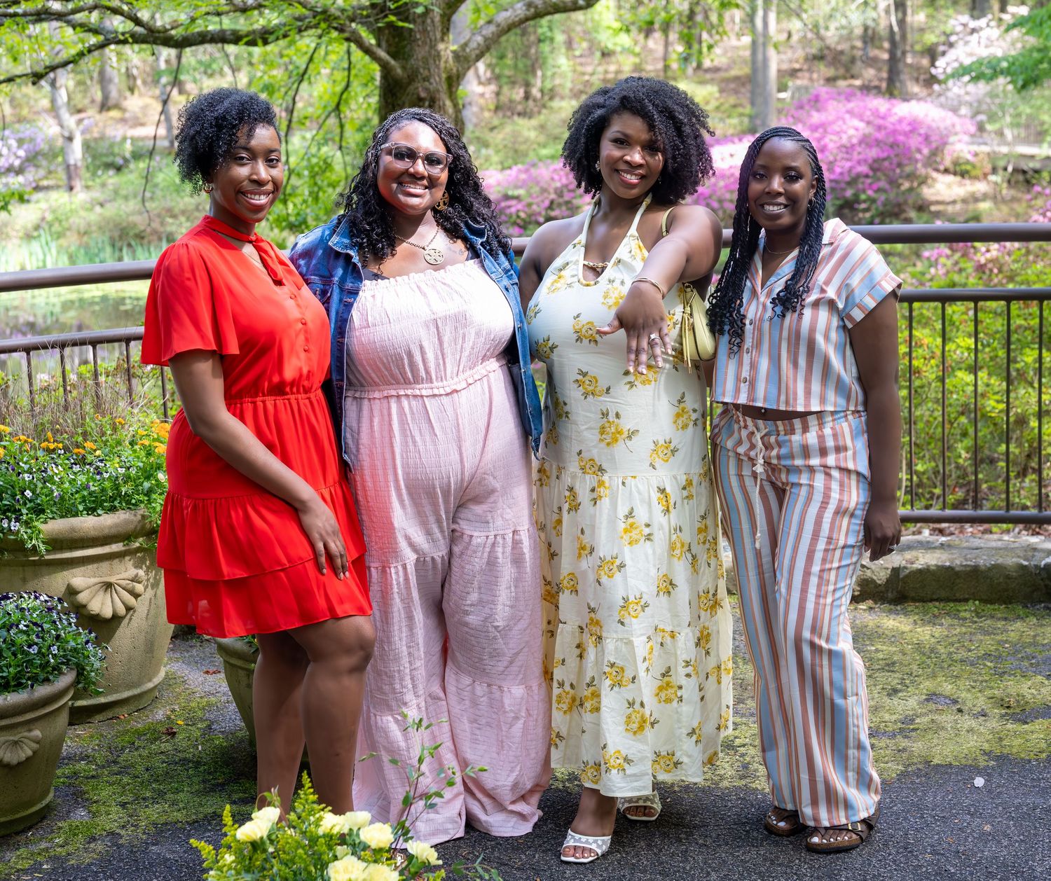 Four women pose together smiling in a vibrant garden with blooming azaleas and colorful flowers in the background.