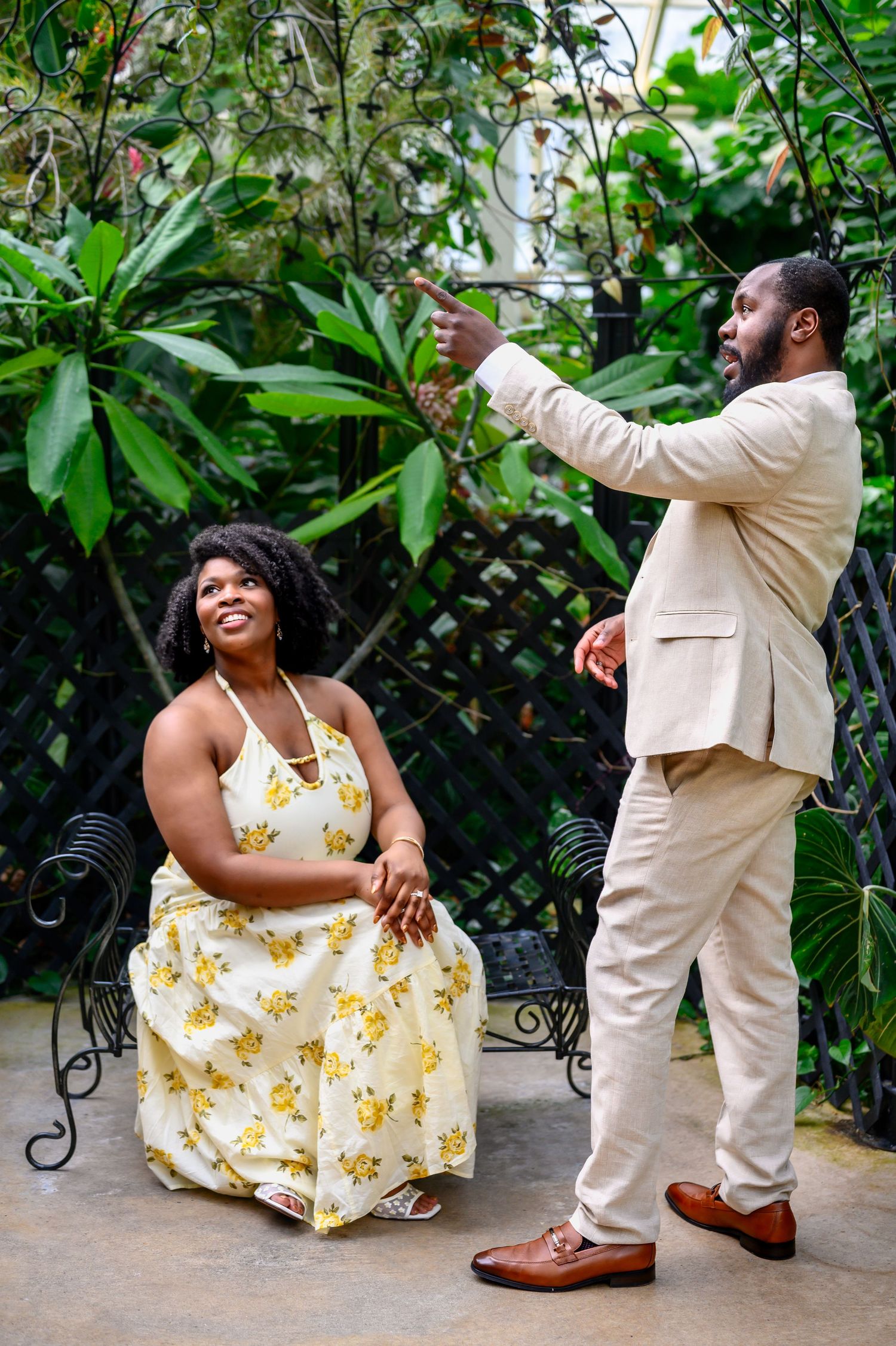 A man in a beige suit points while a woman in a yellow floral dress sits smiling outdoors.