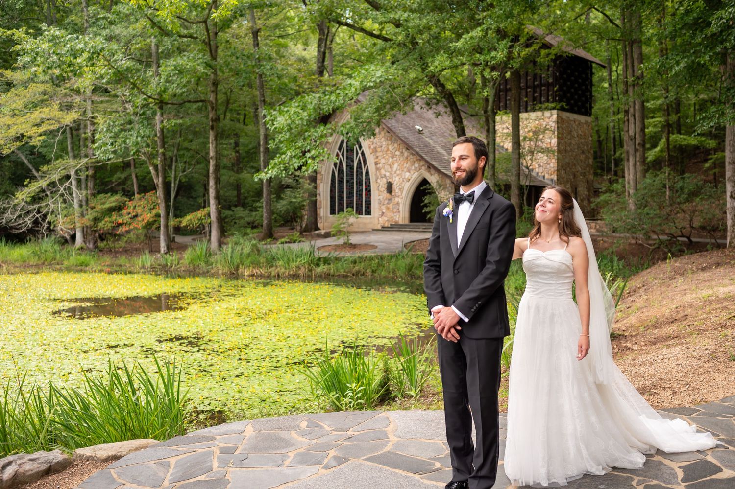 A couple in formal wedding attire strolls through a lush wooded area on a sunny afternoon.