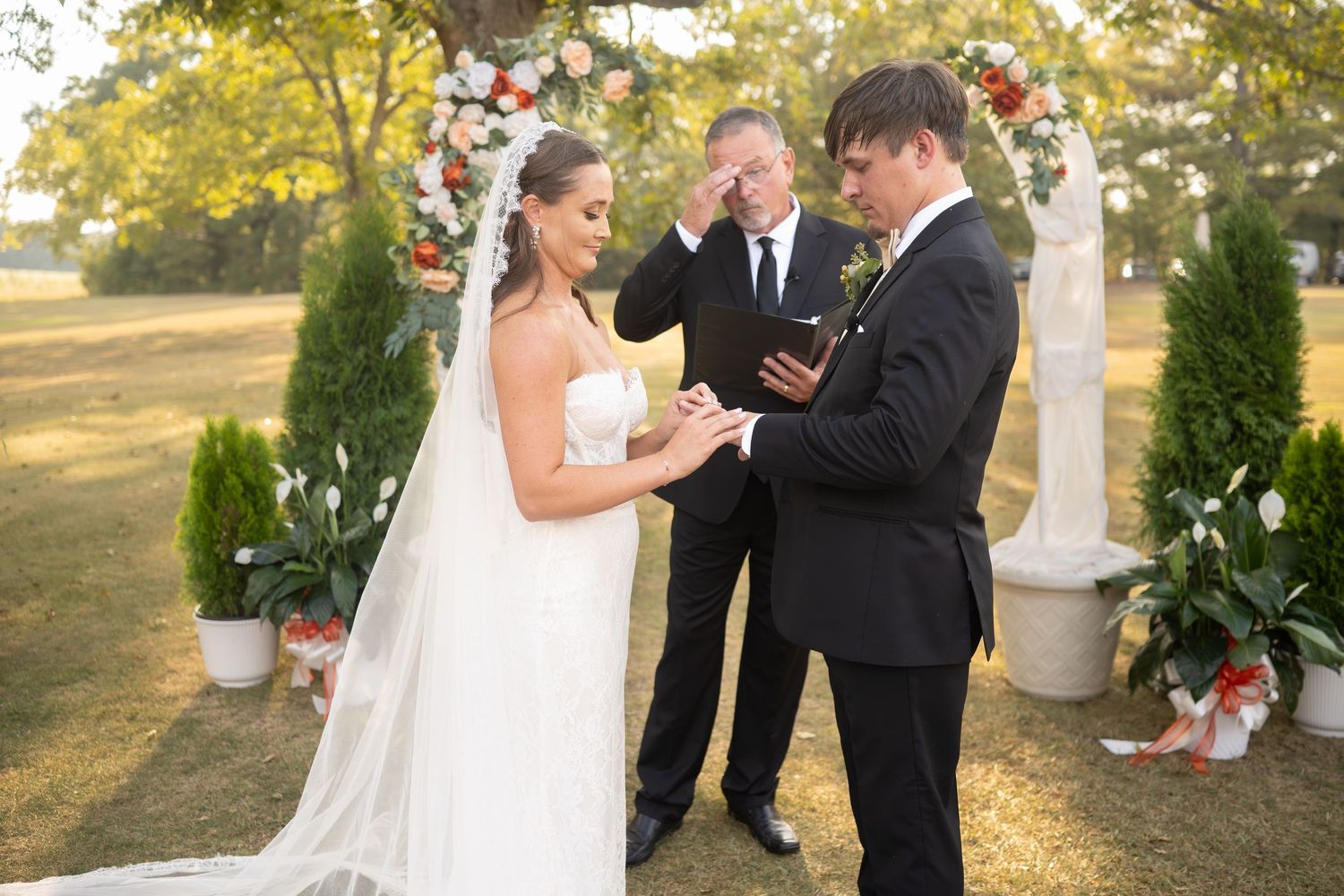 Outdoor wedding ceremony with couple exchanging rings in front of lush greenery and trees.