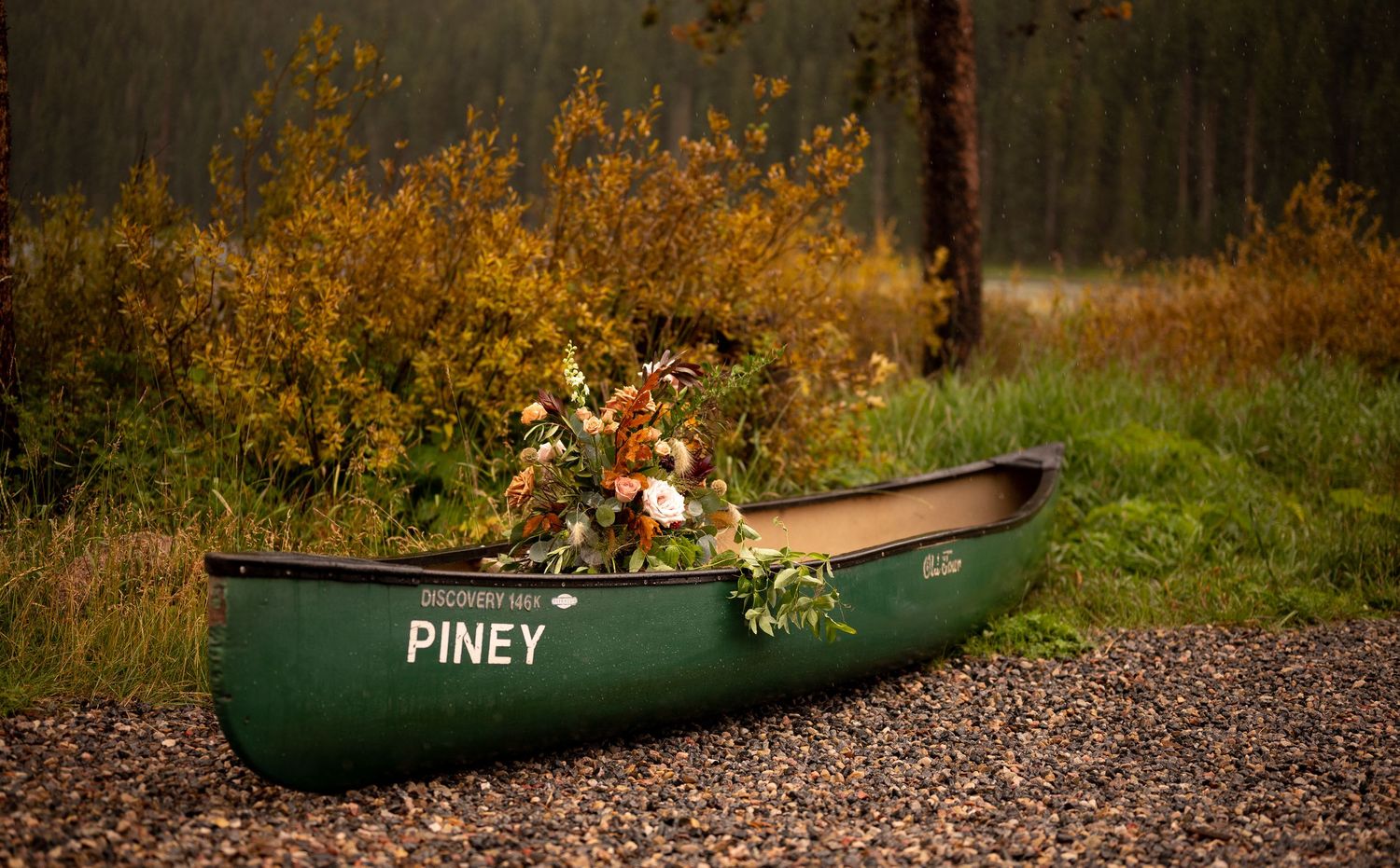 Green canoe named Piney resting on rocky ground surrounded by autumn foliage.