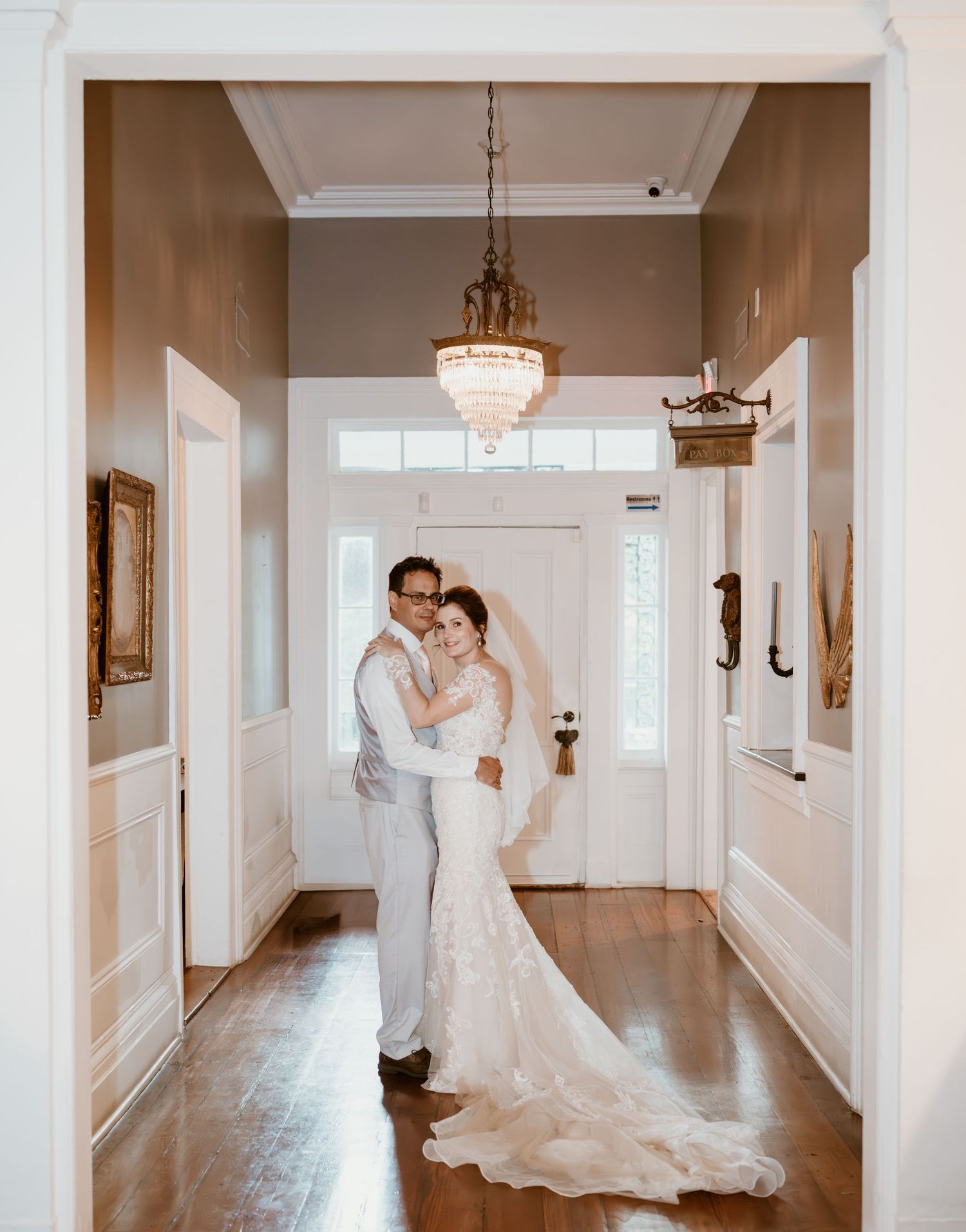 Two people share an intimate moment in a sunlit hallway of an elegant venue with white walls and hardwood floors.