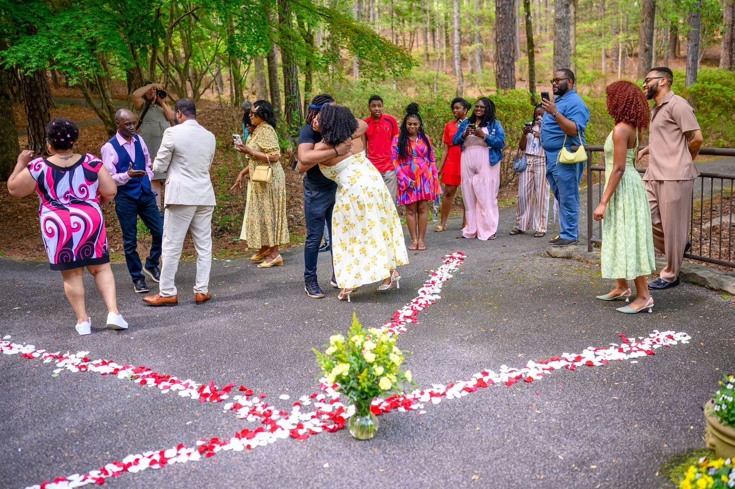 Newly engaged couple kissing while surrounded by celebrating family and friends on rose petal decorated path.