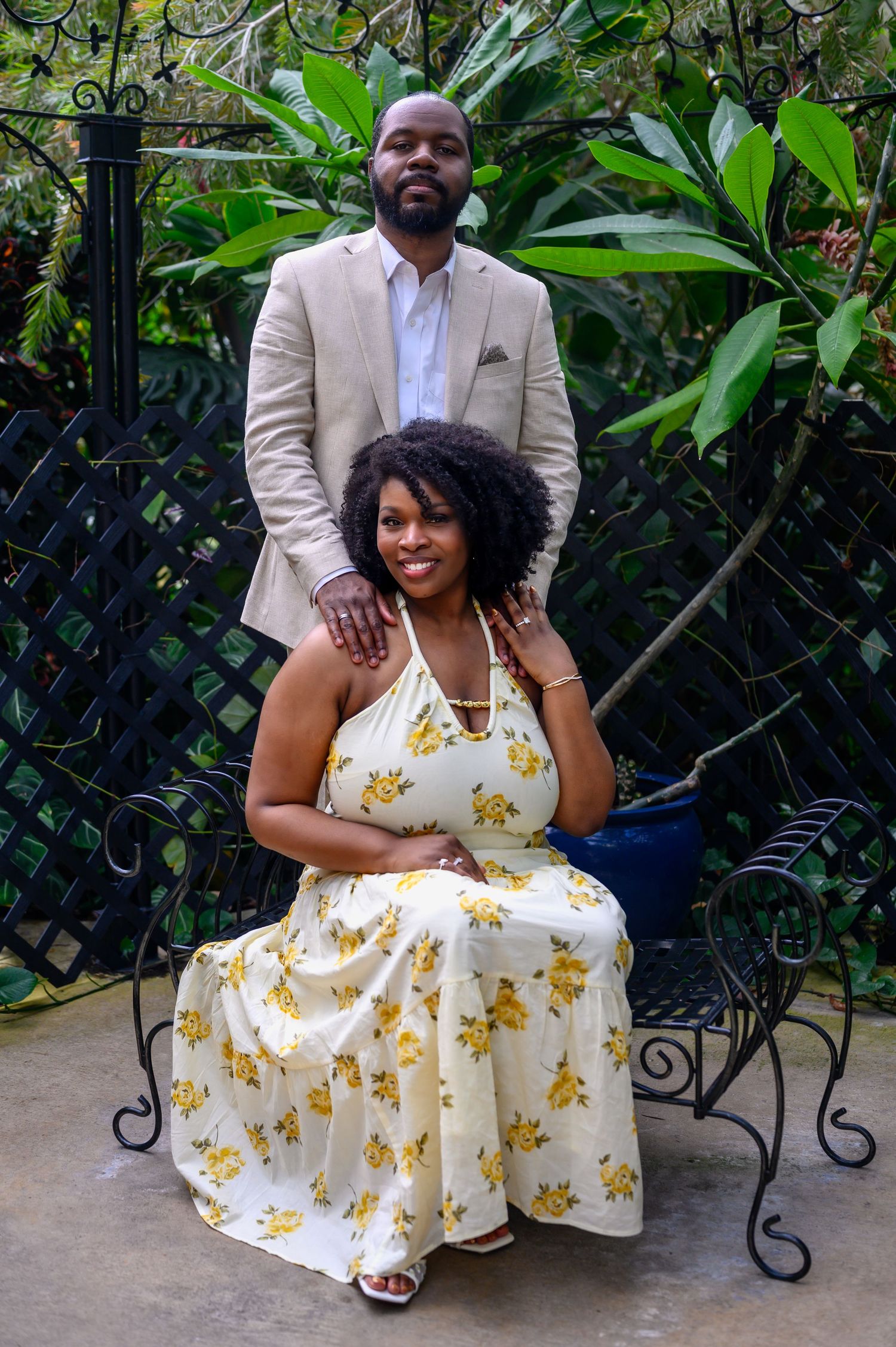 A couple poses in a lush garden, woman in floral dress seated on iron bench, man in tan blazer standing behind her.