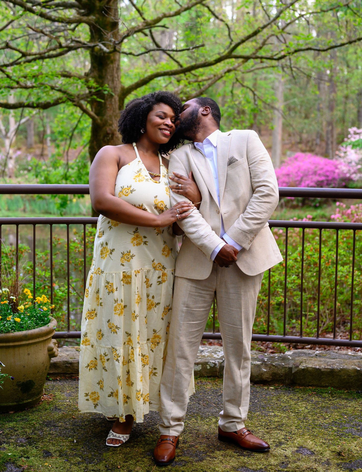 A couple shares an intimate moment outdoors, the man kissing her cheek near blooming azalea bushes.