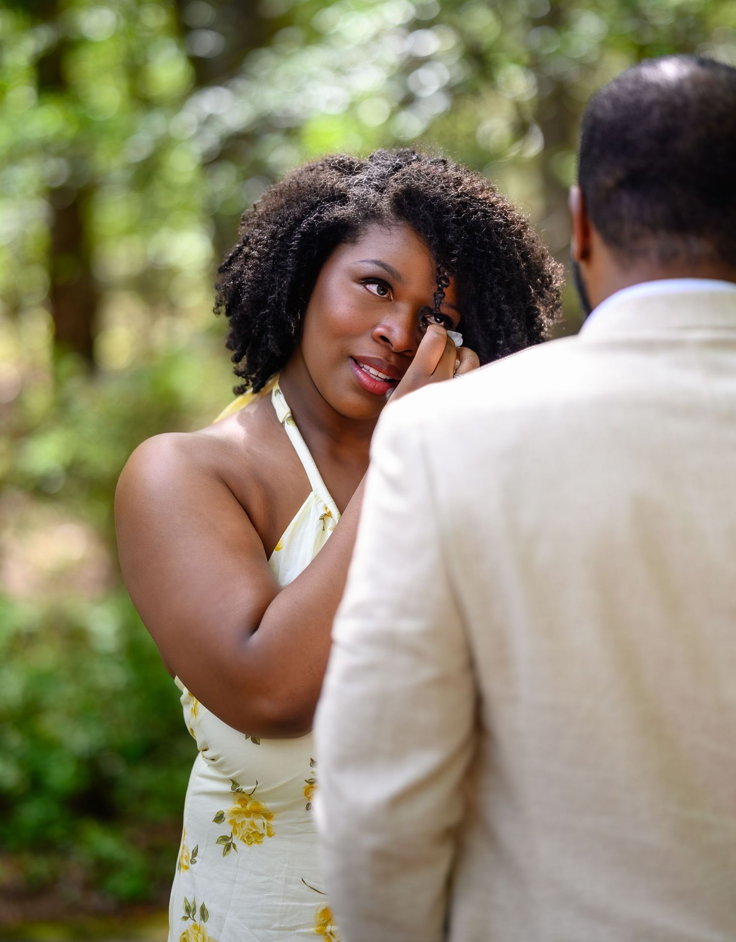 Emotional woman in yellow floral dress wiping tears during romantic outdoor marriage proposal in wooded garden.