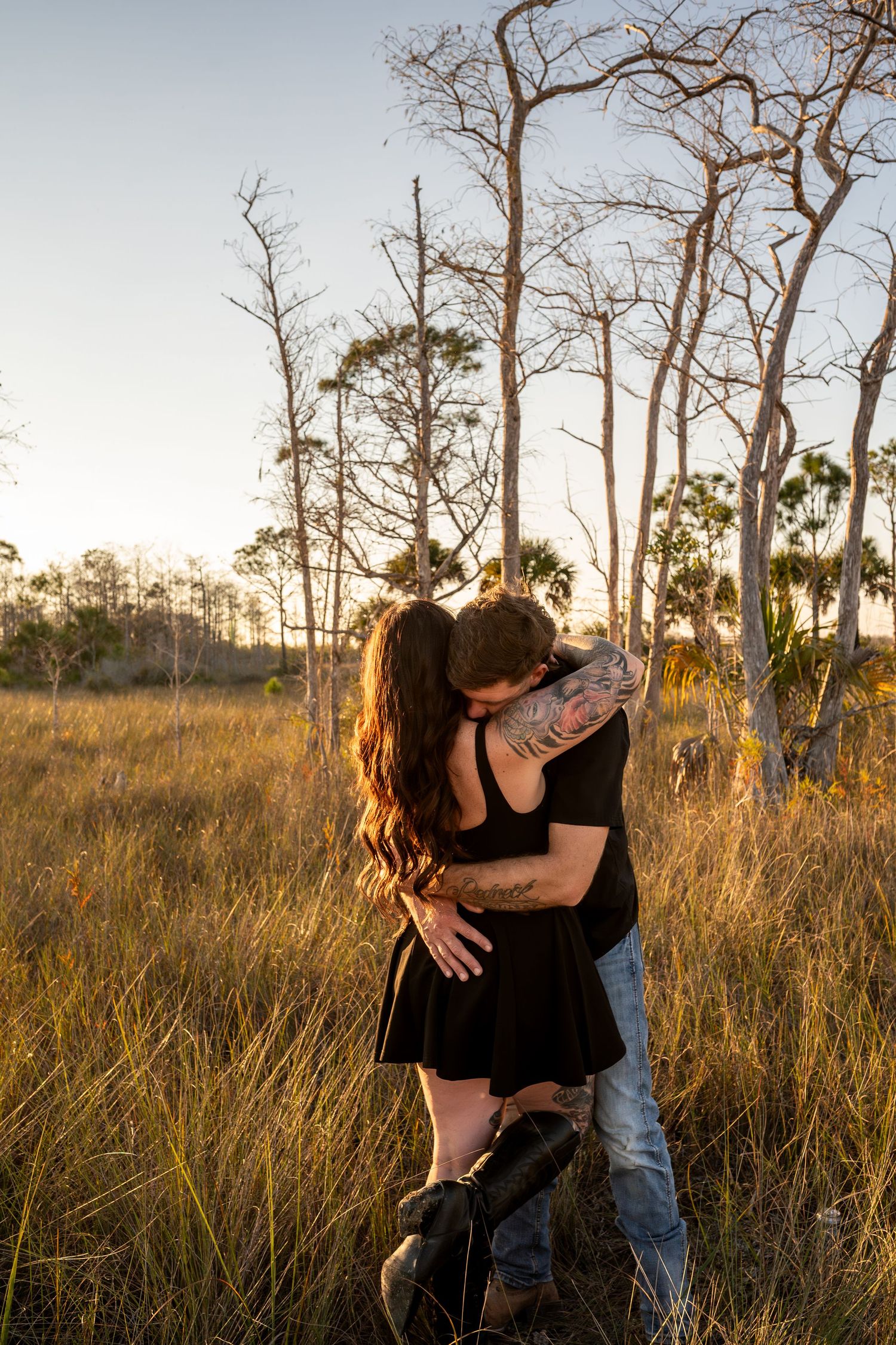 Couple hugging and laughing together in a field with bare trees at sunset.