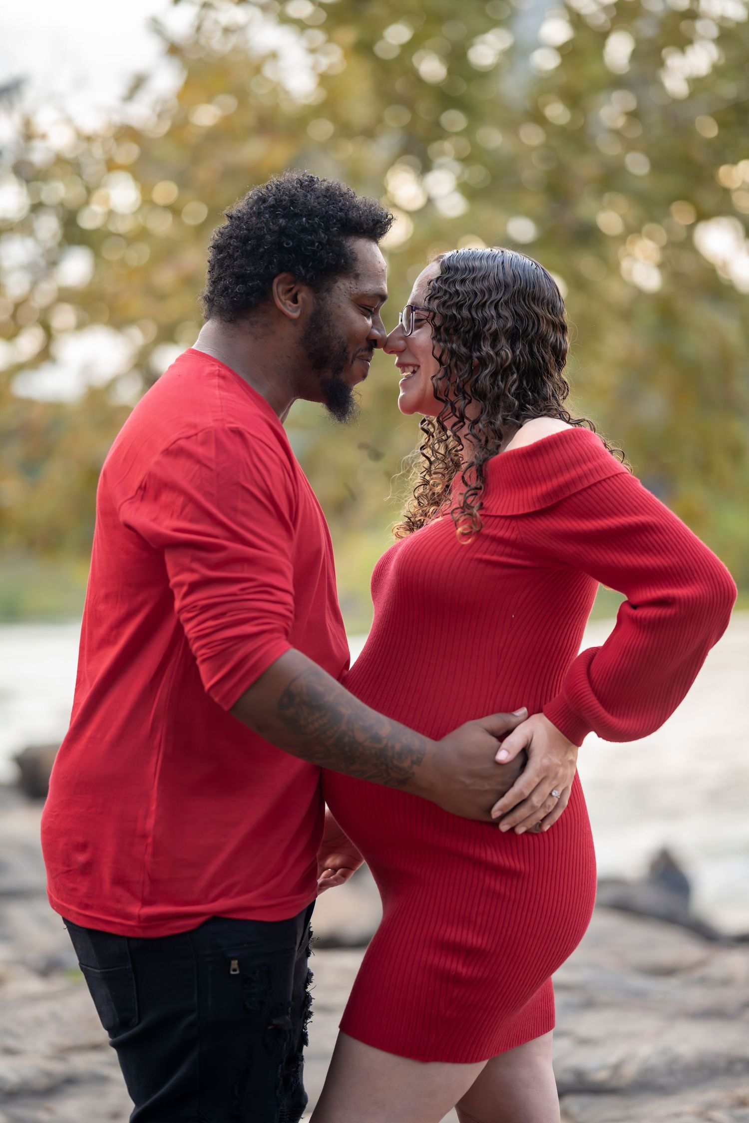 Couple wearing red embraces on rocky shore during autumn maternity photo session.