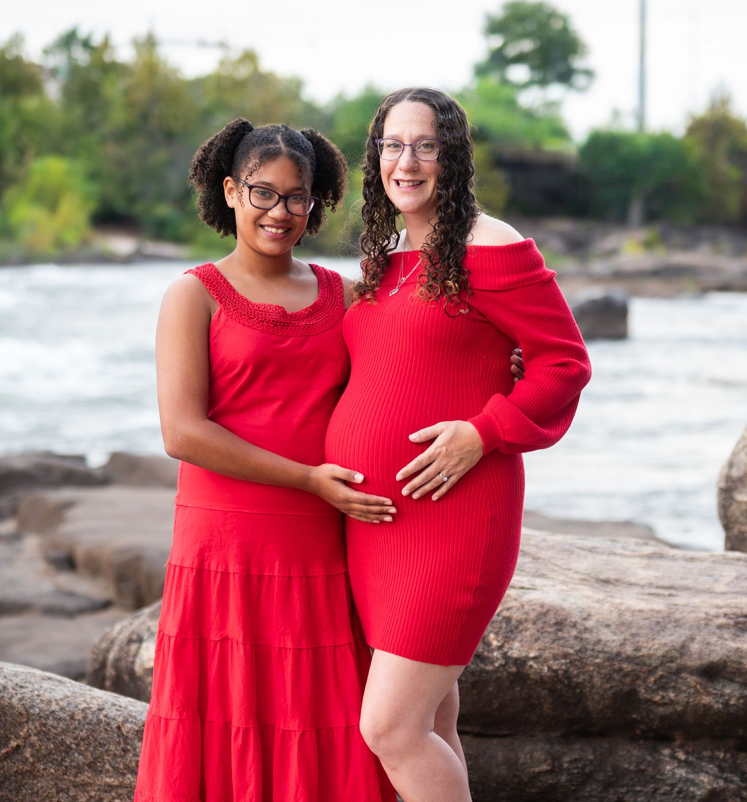 People in matching red dresses stand together on rocky shoreline with water and trees in background.