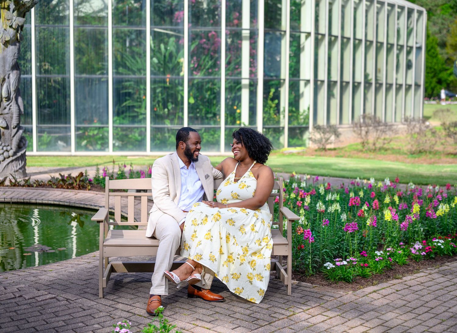 Engaged couple sitting on a bench beside a pond with tulips and a greenhouse.