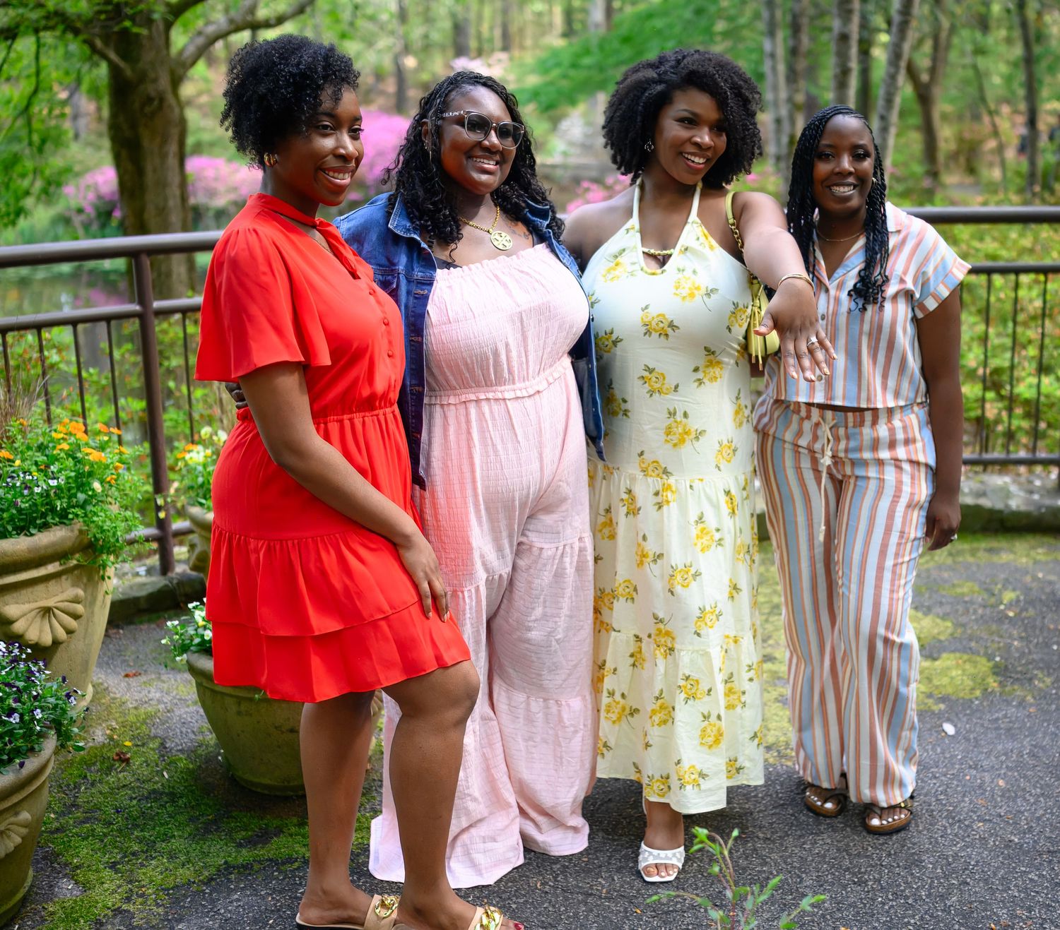 Four women pose together smiling in a garden with colorful flowers and greenery in the background.