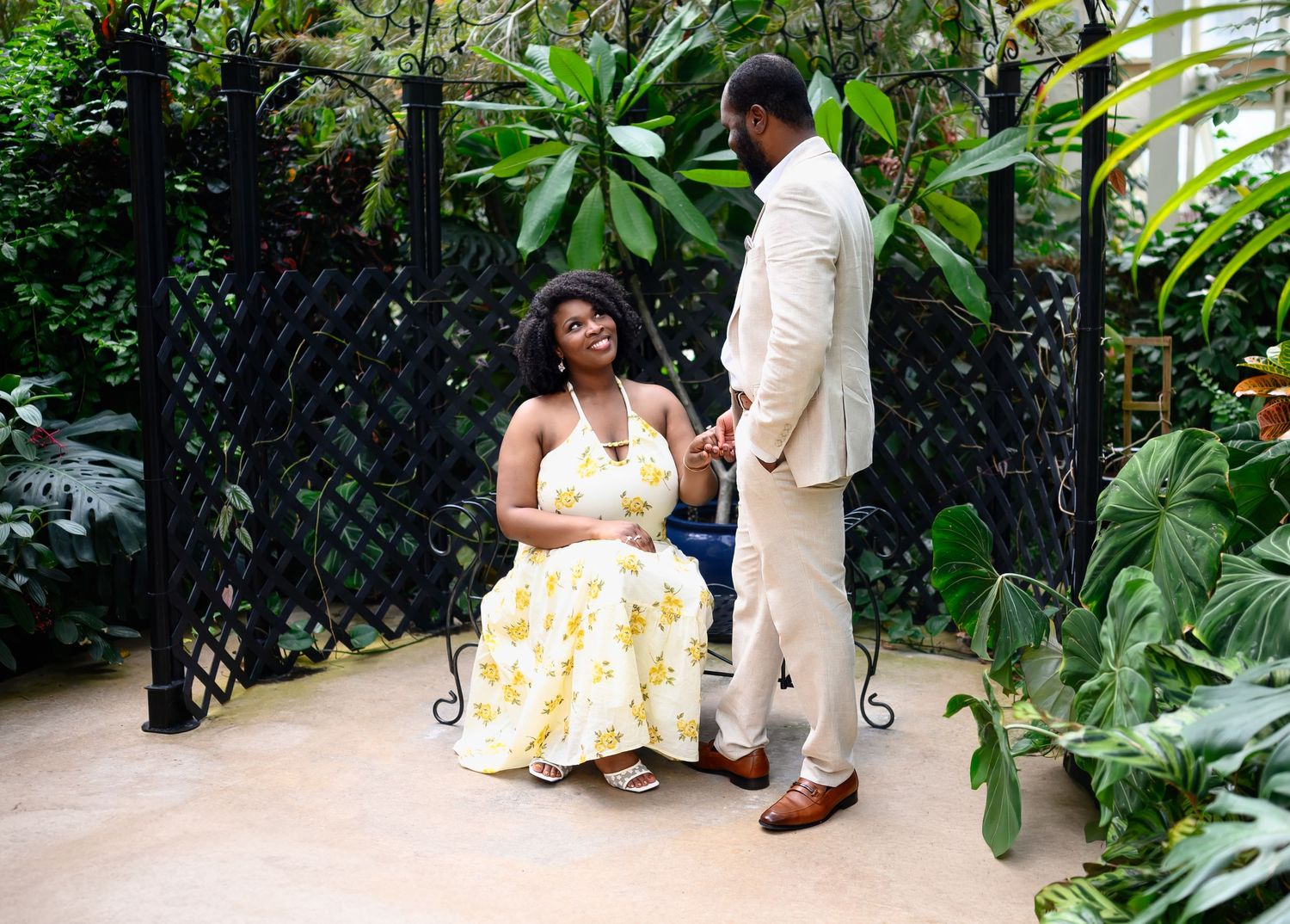 Couple sharing a tender moment seated among lush tropical plants and black iron gates.