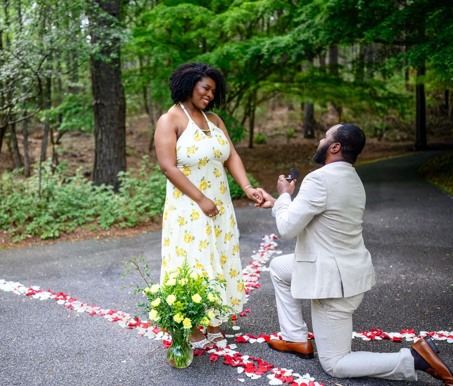 Man kneels on one knee proposing to woman in yellow floral dress on a petal-decorated forest path.