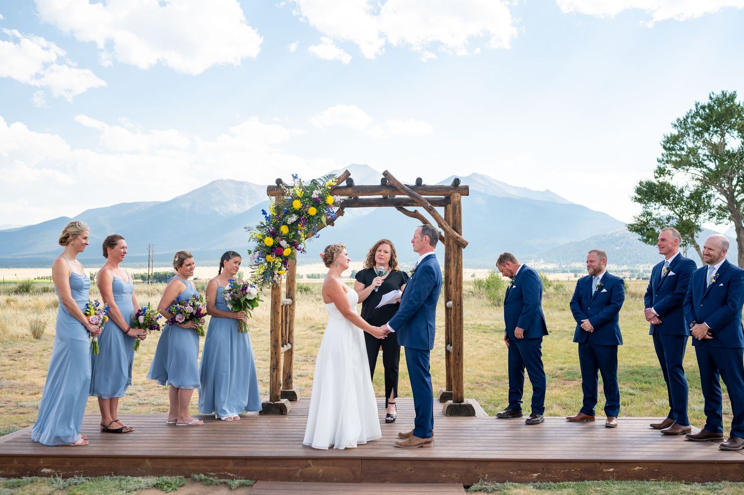Wedding party in blue attire posed on wooden deck with mountain range in distance at the wedding venue, Barn at Sunset Ranch.