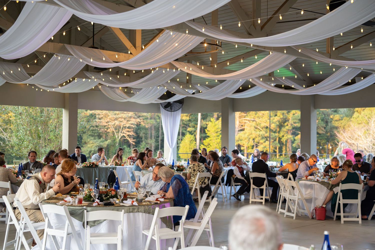 Wedding reception venue with white draping and string lights overhead, tables set for dining.