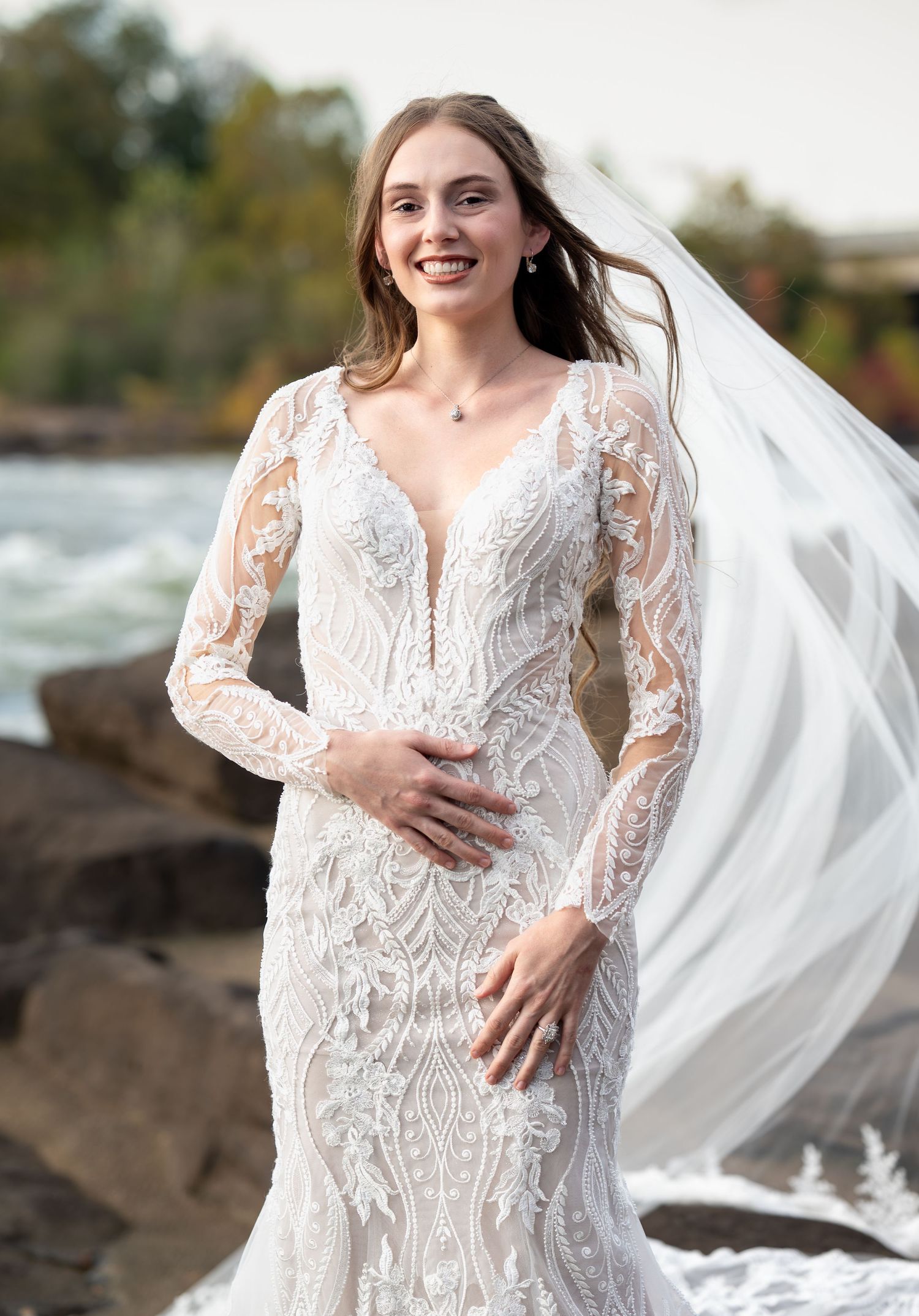 Bride in elegant lace long sleeve wedding dress poses outdoors near rocky waterfront with flowing veil in autumn.