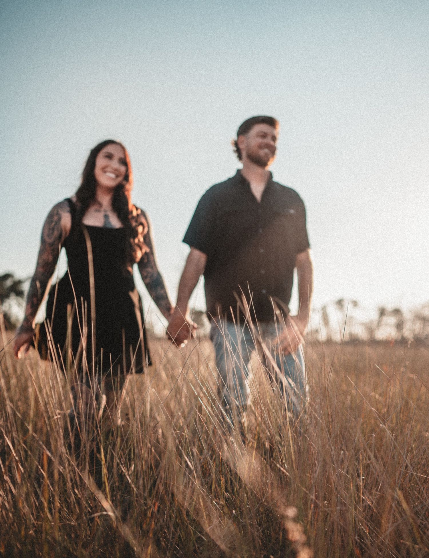Couple walking hand in hand through tall grass during a sunset photoshoot.