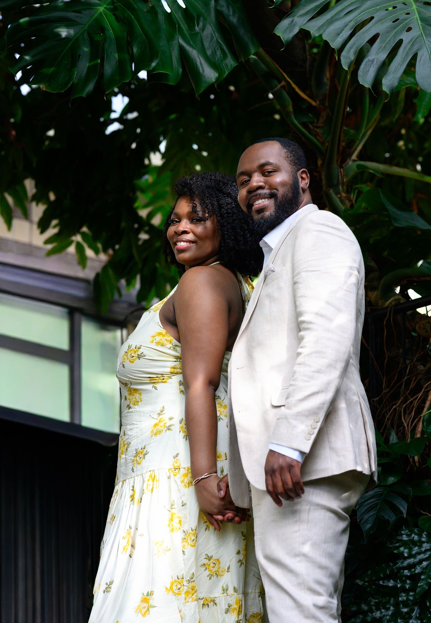 A smiling couple holds hands and poses together in front of large tropical monstera leaves in a garden setting.