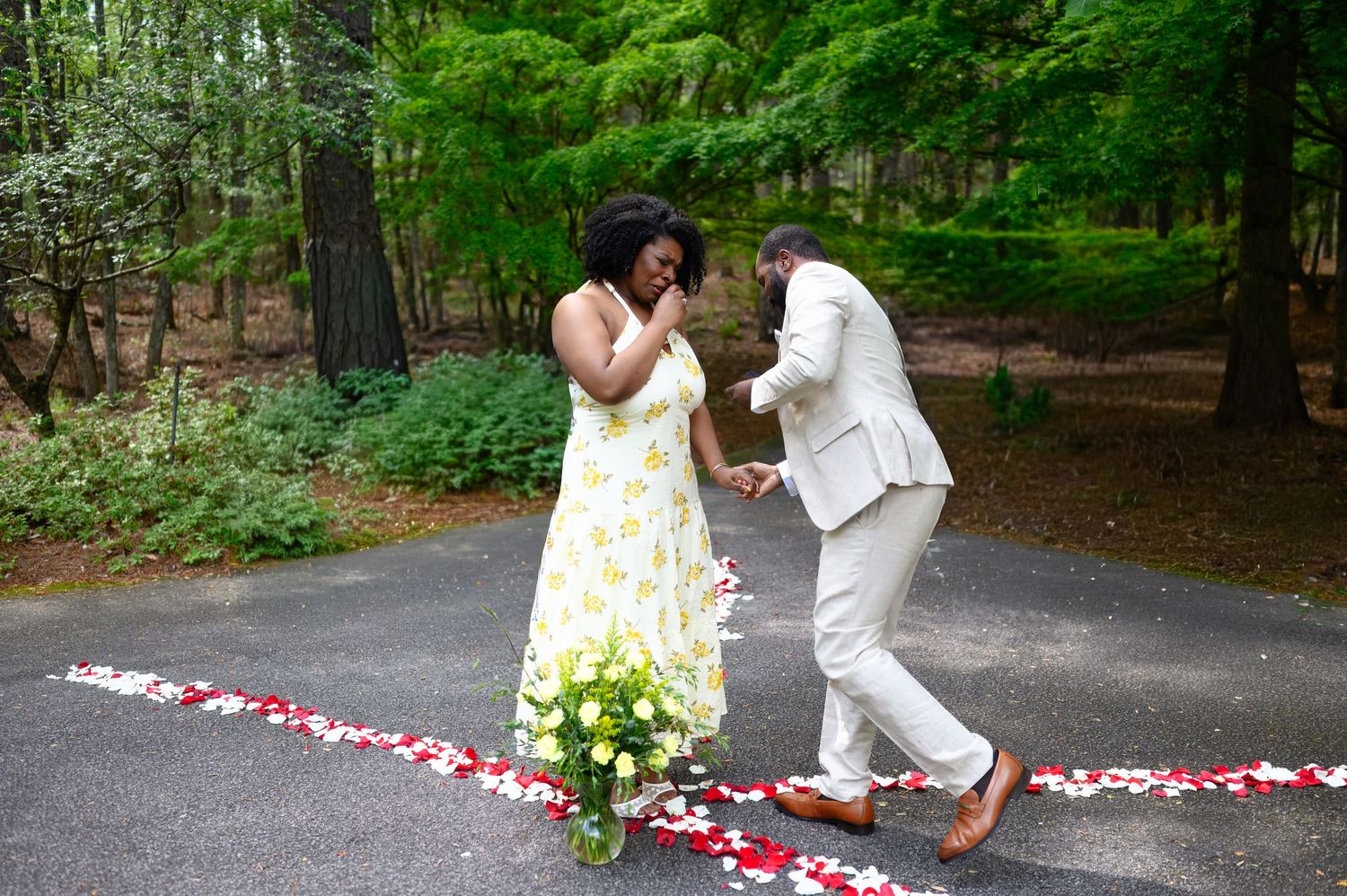 A couple dances joyfully on a petal-lined path surrounded by trees, with white flowers nearby.