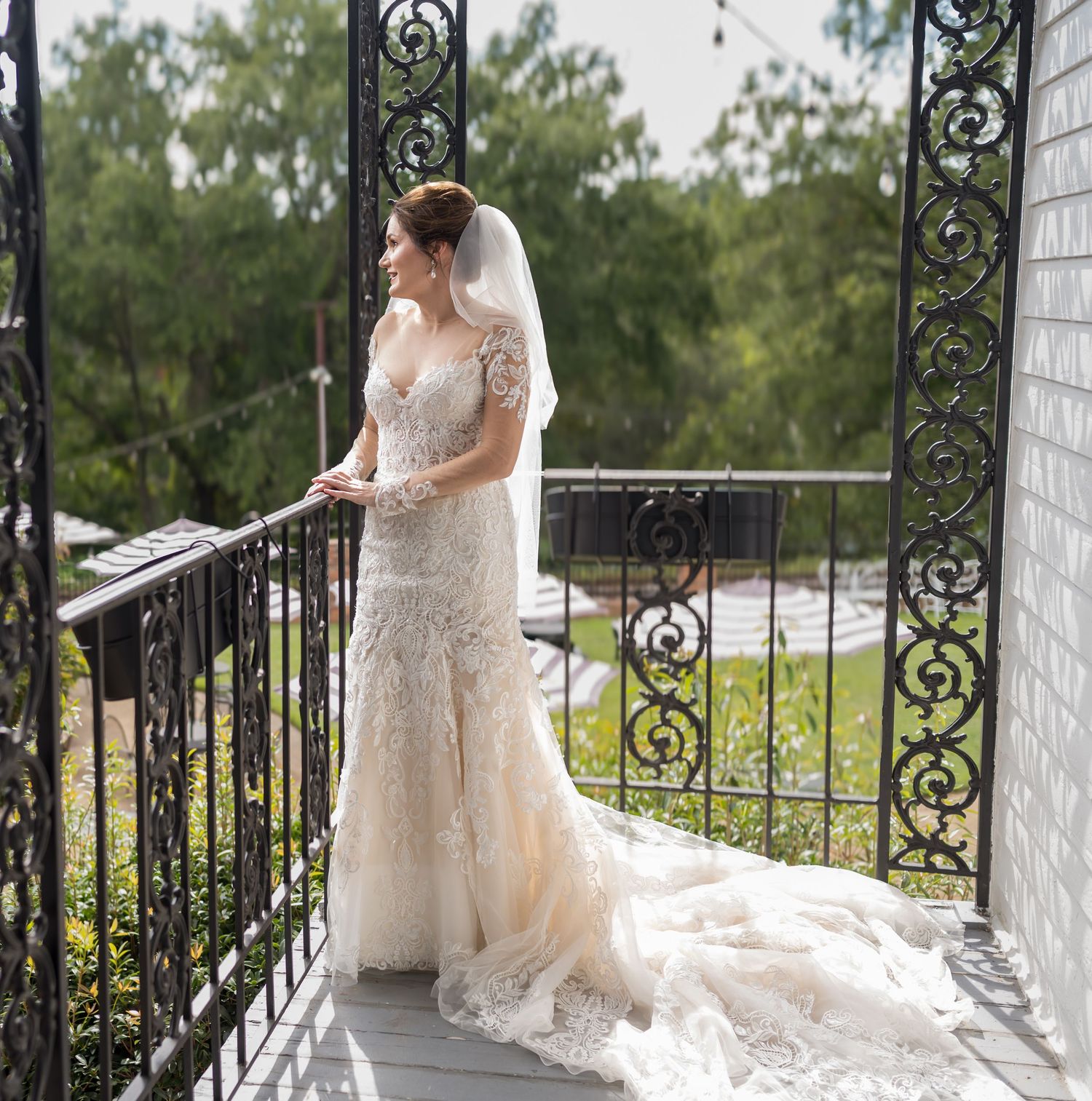 A bride in a flowing lace gown with a long train poses gracefully on a white wraparound porch with ornate black railings.