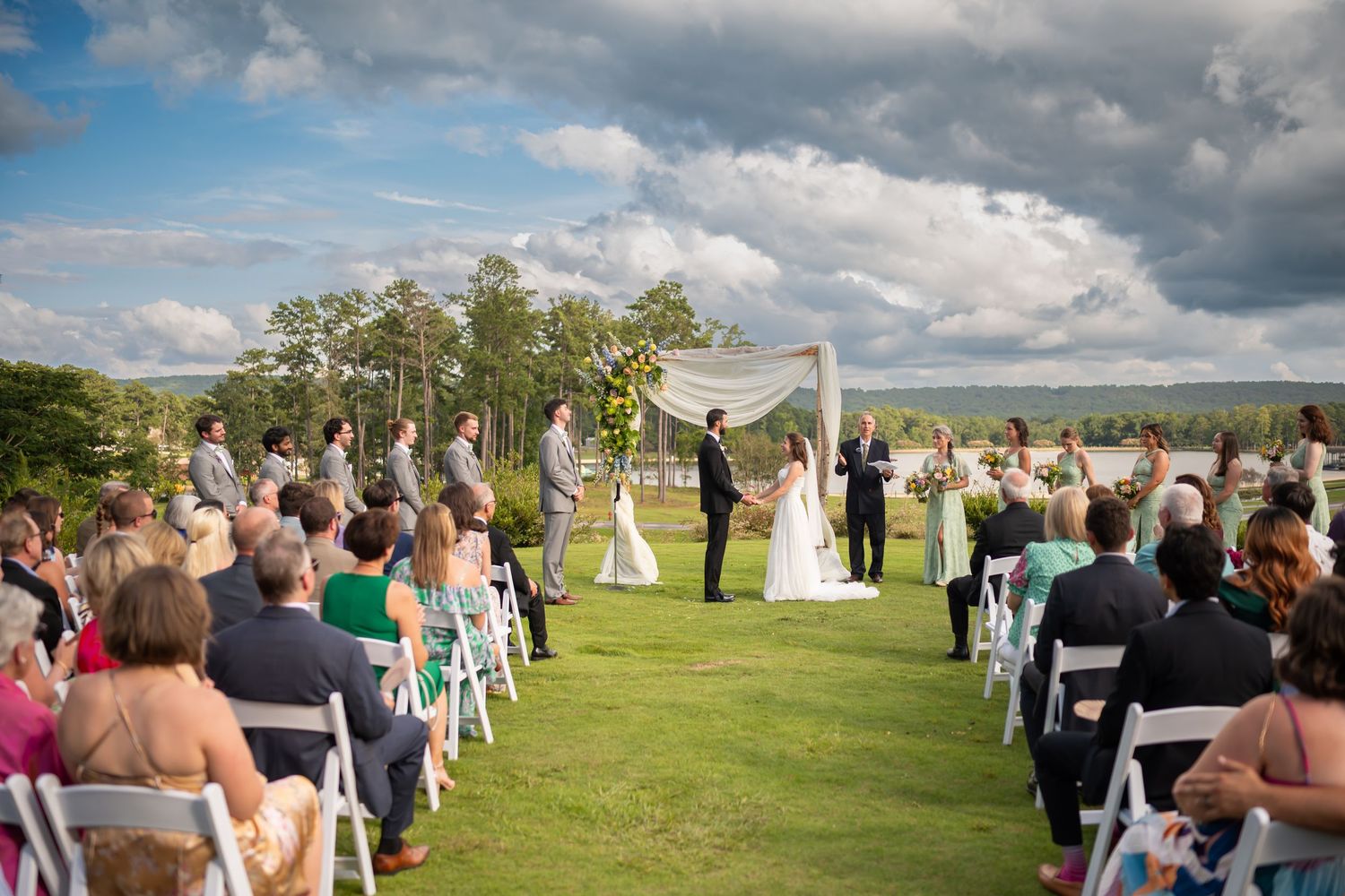Outdoor wedding ceremony on a lush green lawn with white chairs and dramatic cloudy sky backdrop.