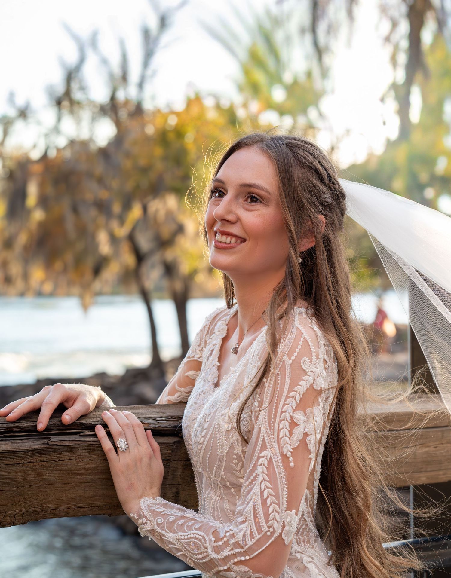 Romantic waterfront bridal portraits featuring a detailed lace gown and flowing veil at sunset.