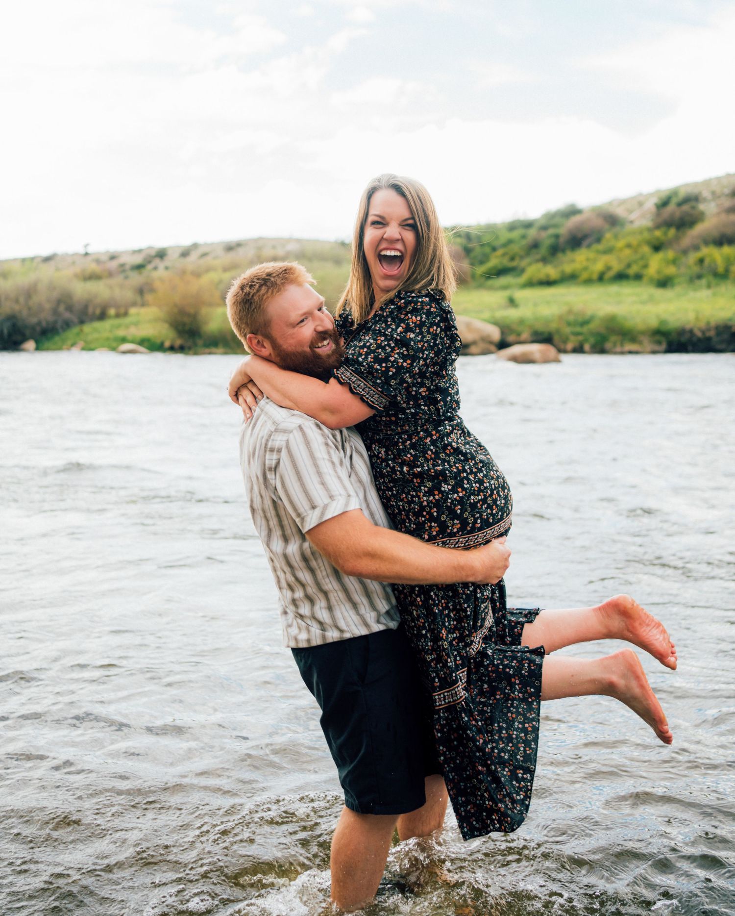 Two people playfully embrace in a lake with mountains in the background.