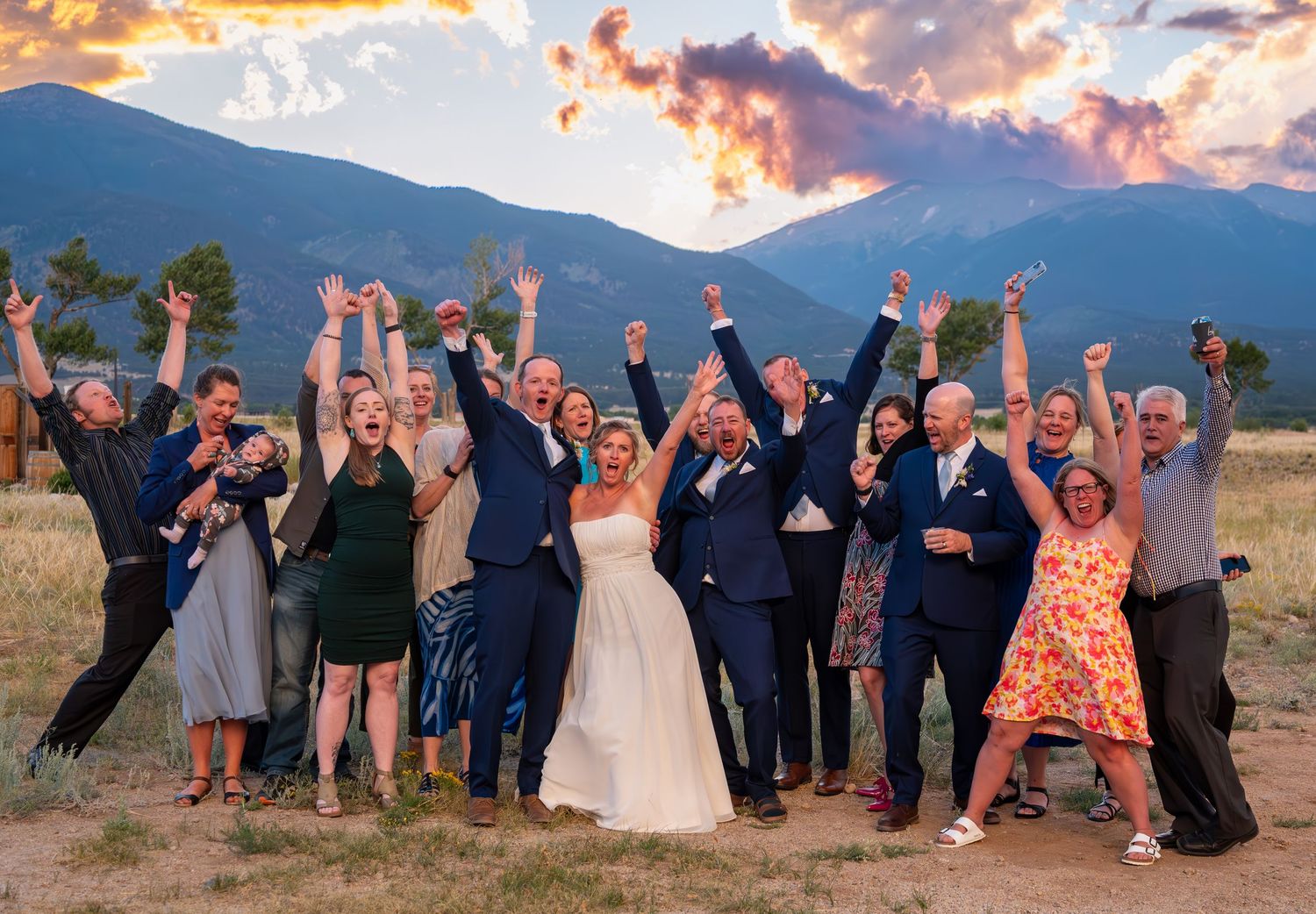 Wedding party celebrating with raised arms against a mountain backdrop at sunset.