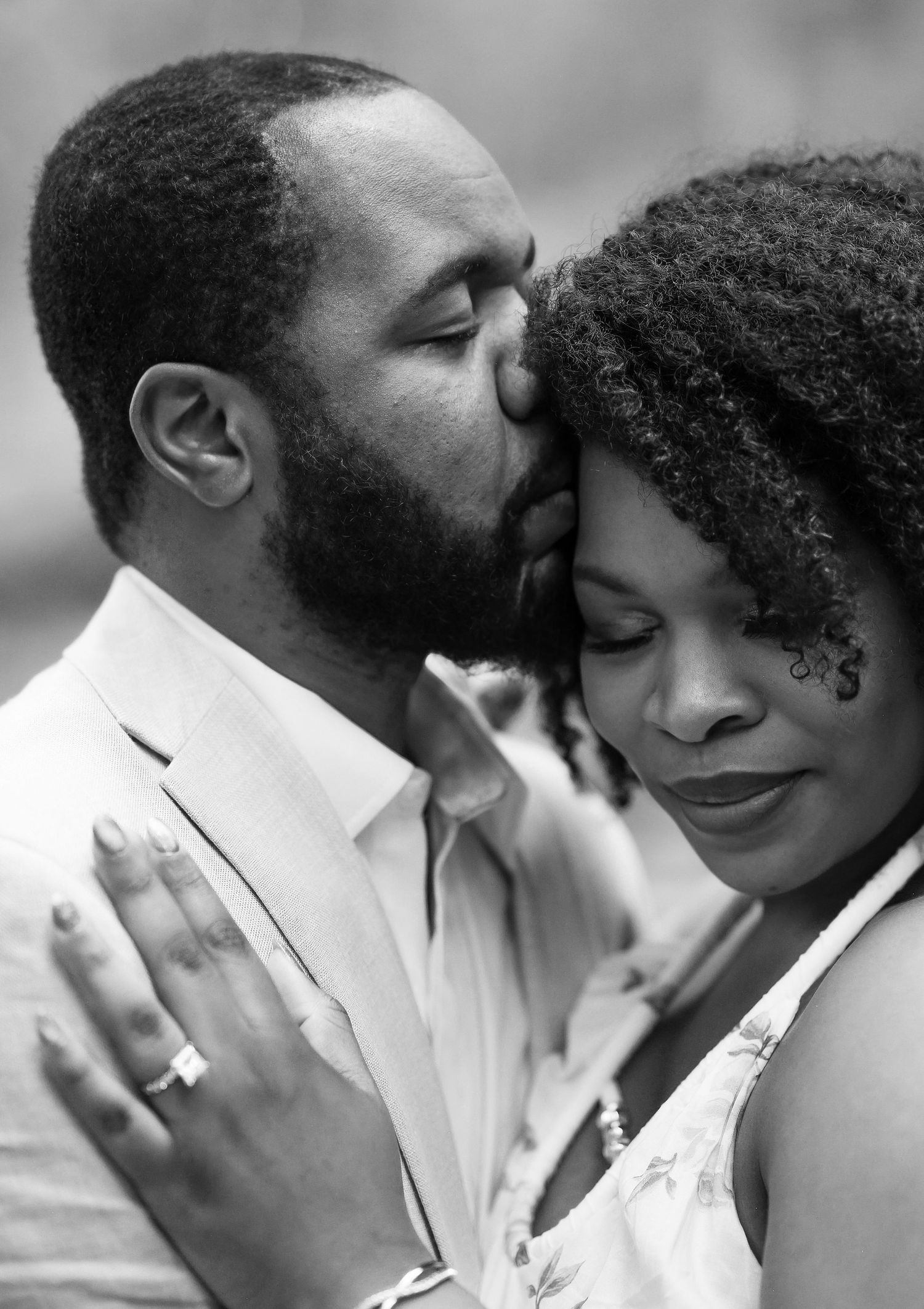 Intimate black and white close-up of a bearded man kissing a woman's forehead, engagement ring visible.