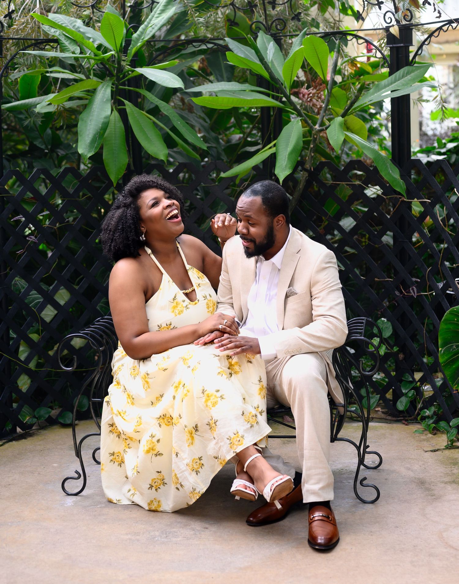 Happy couple laughing together while sitting on an ornate black iron bench surrounded by tropical greenery.
