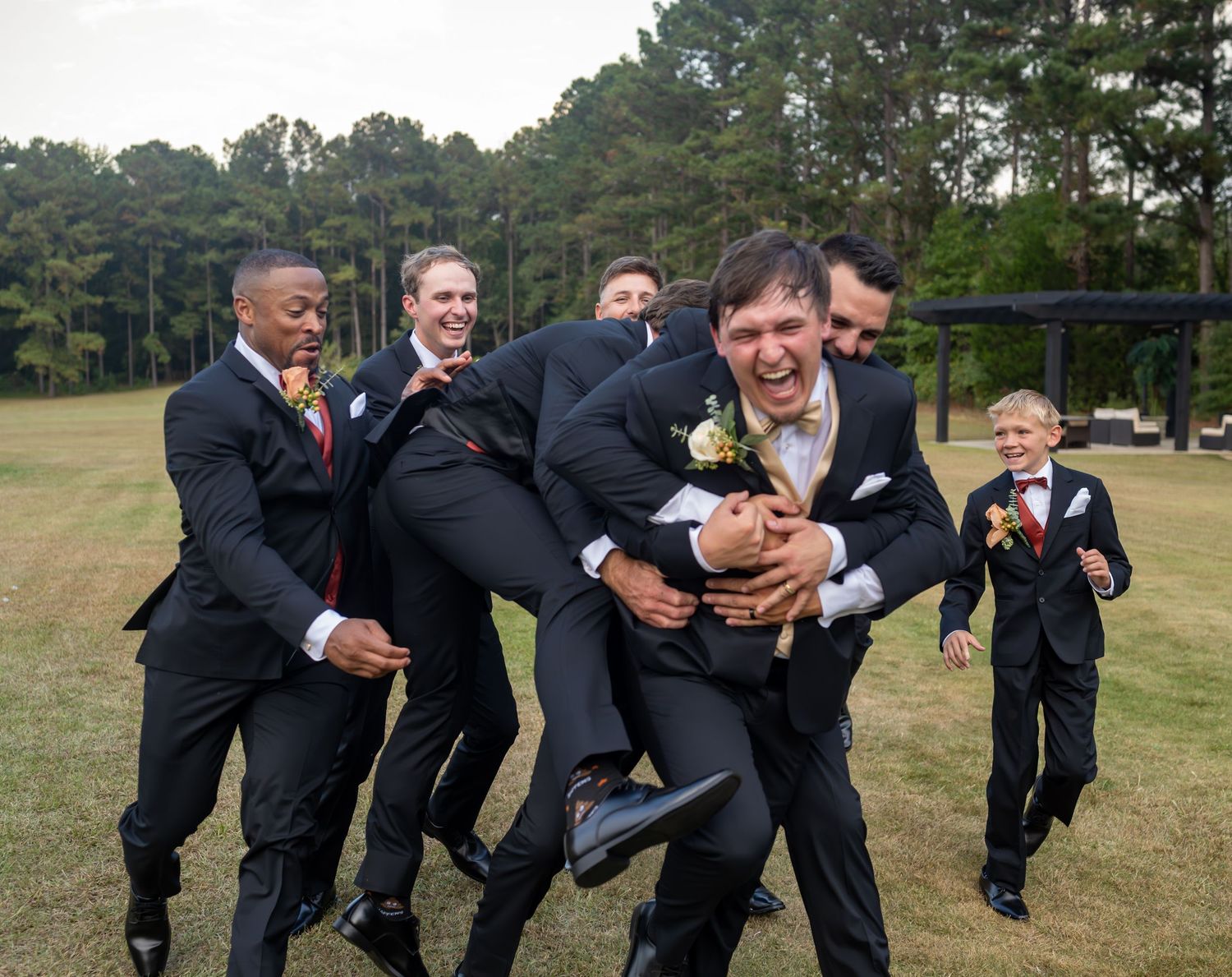 Groomsmen in black suits playfully posing together on grassy field.