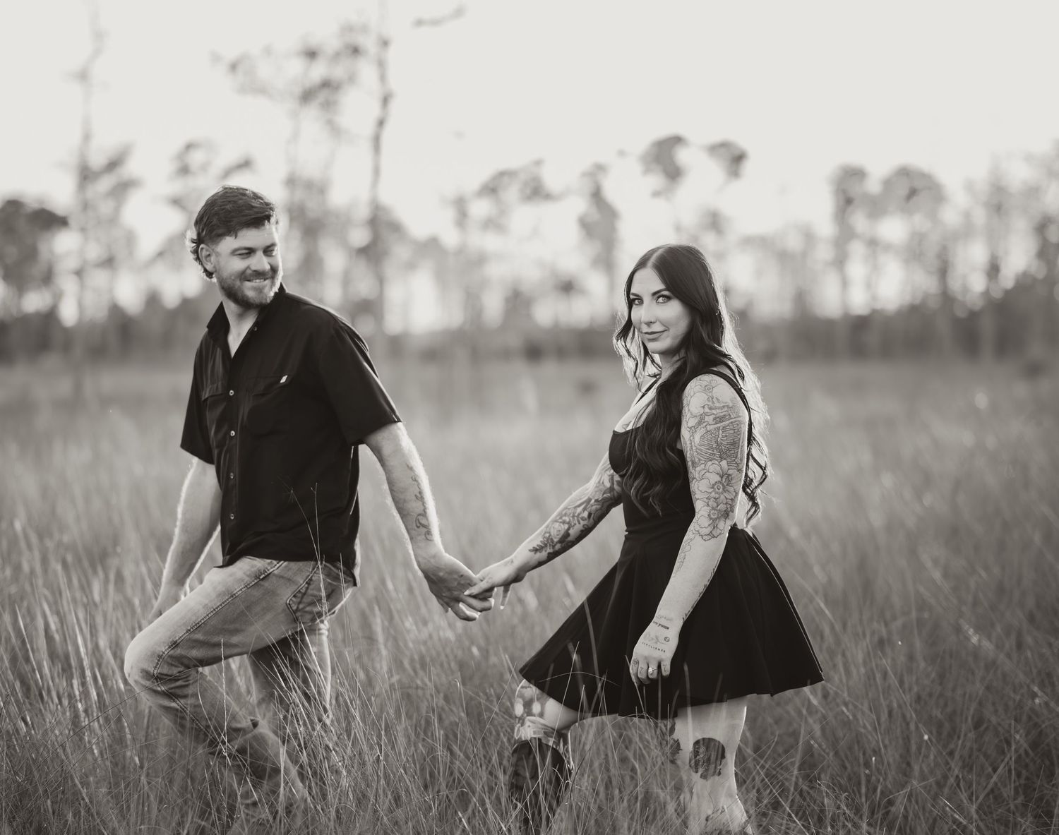 A couple in black clothing holds hands and walks through a tall grass field in a romantic black and white photograph.