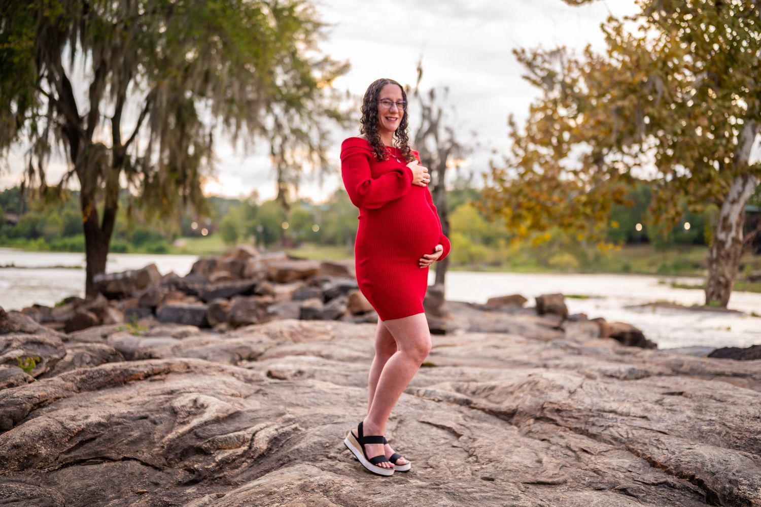 Person in a vibrant red dress poses on natural rock formations near a lake with autumn trees in background.