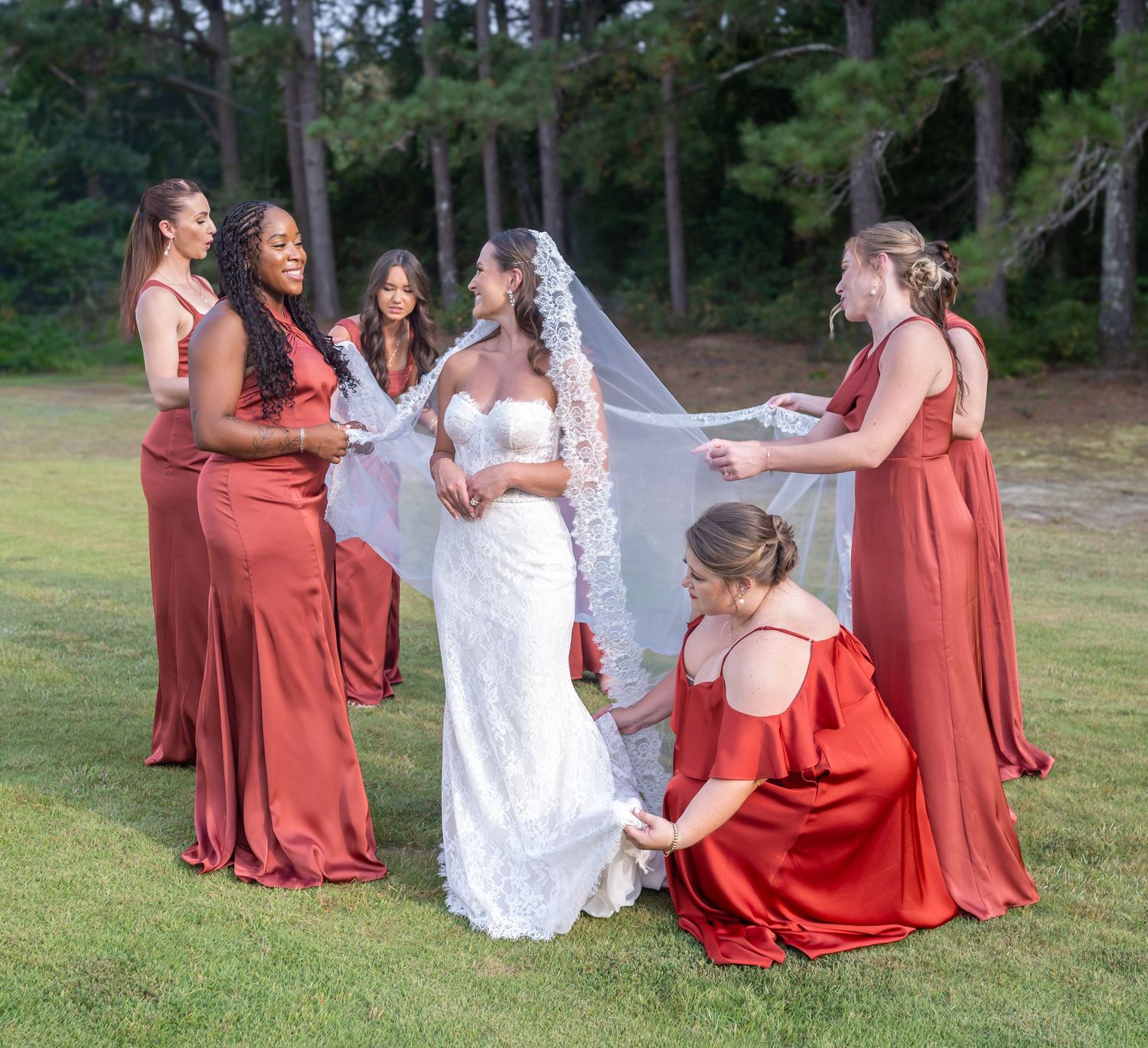 Bridesmaids in flowing rust-colored dresses gather around a bride in white lace on a grassy lawn.
