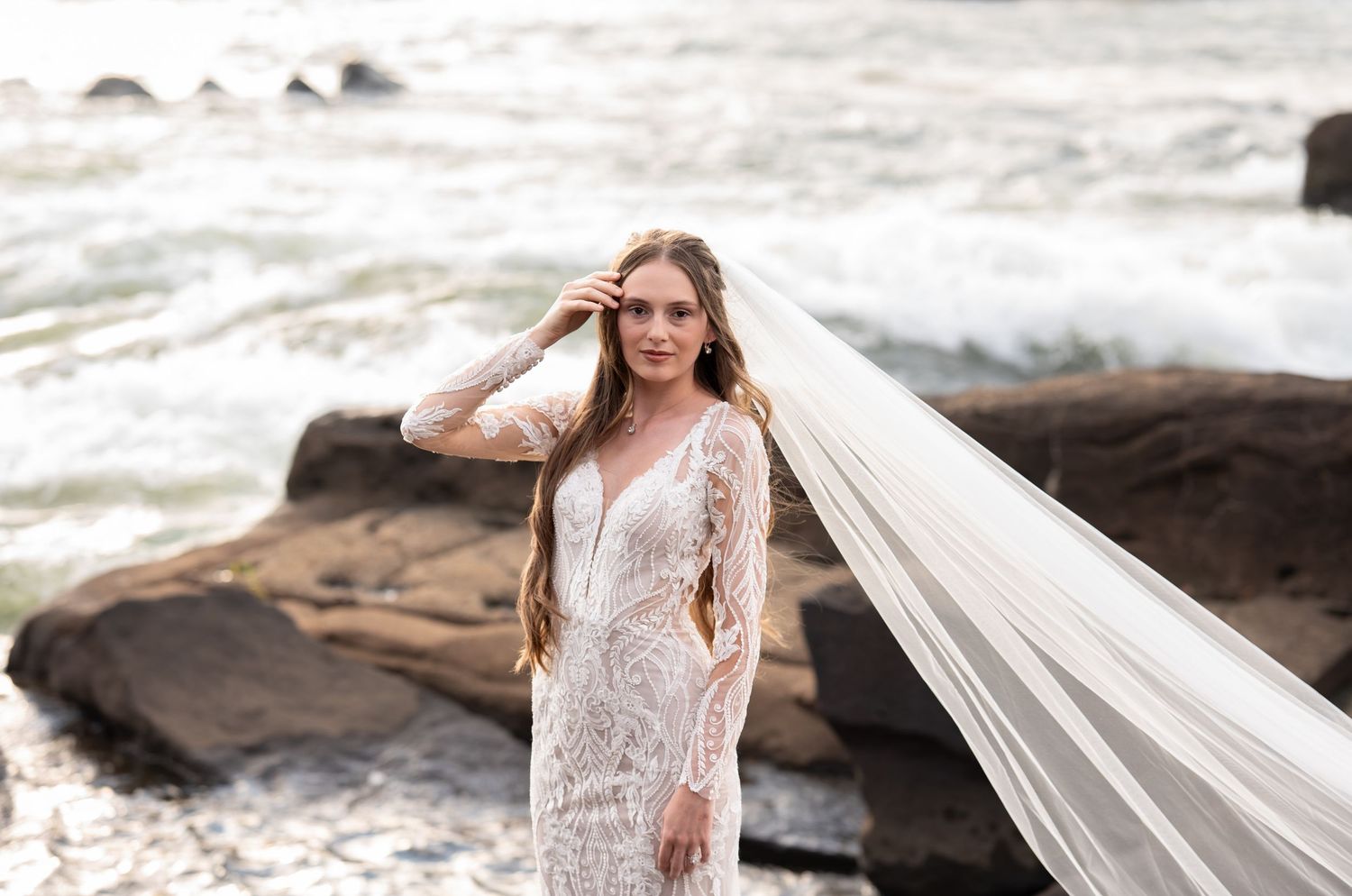 A bride in a flowing white dress poses gracefully on a rocky beach with her veil floating in the ocean breeze.