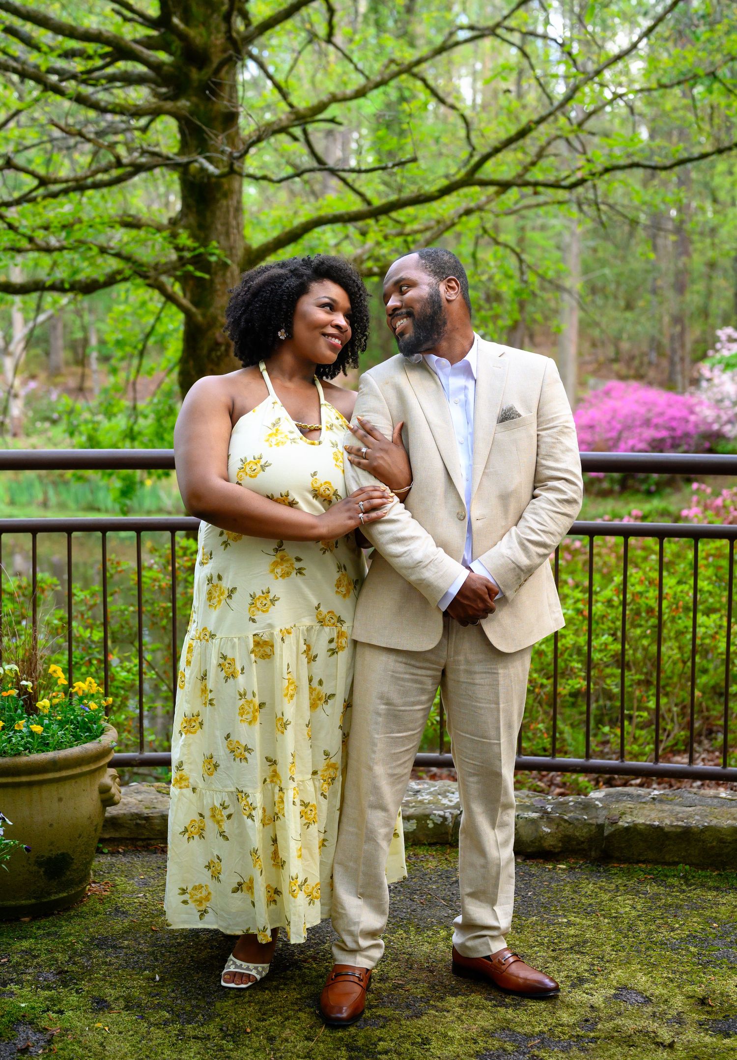 A couple smiles at each other in a lush garden, woman in floral dress, man in tan suit, spring blooms behind them.