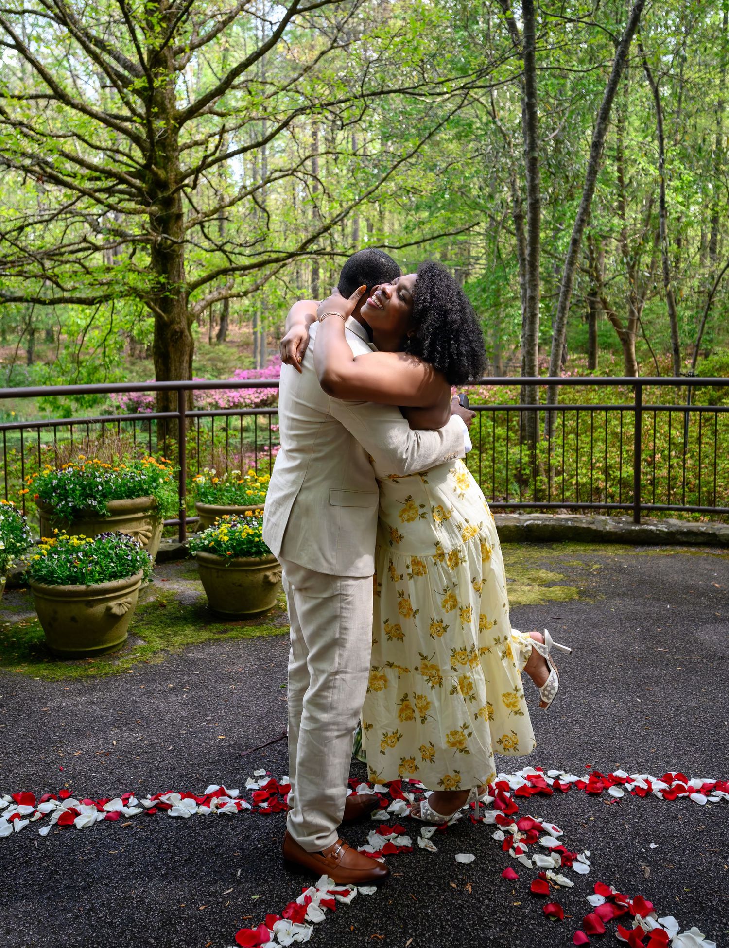 Newly engaged couple shares a joyful embrace on rose petal-decorated patio surrounded by spring blooms.